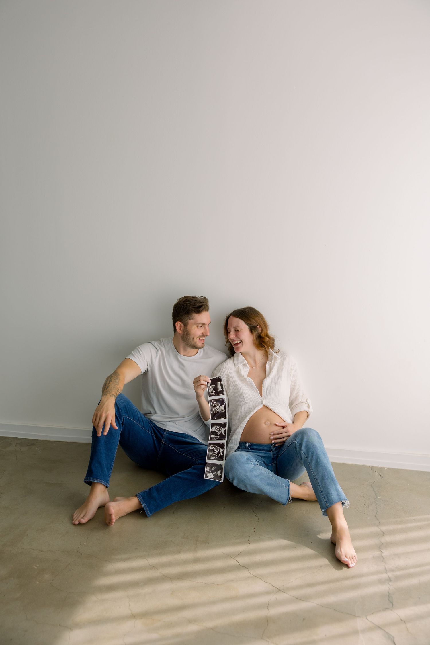 Happy expecting couple sitting on floor holding ultrasound photos next to pregnant belly.