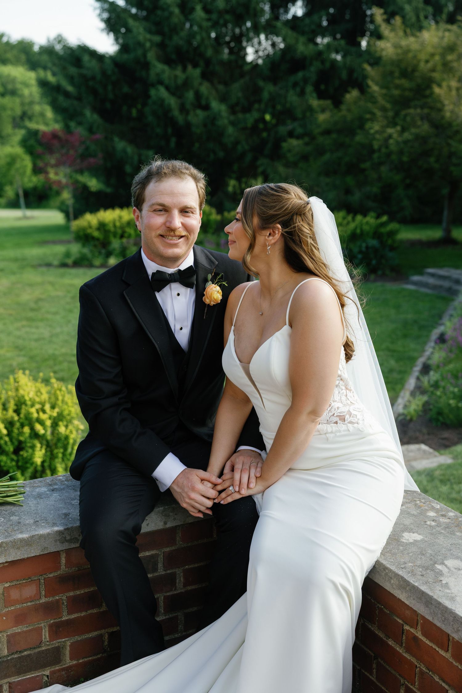 Bride and groom sitting on a brick wall holding hands in a lush garden during their outdoor wedding portrait.