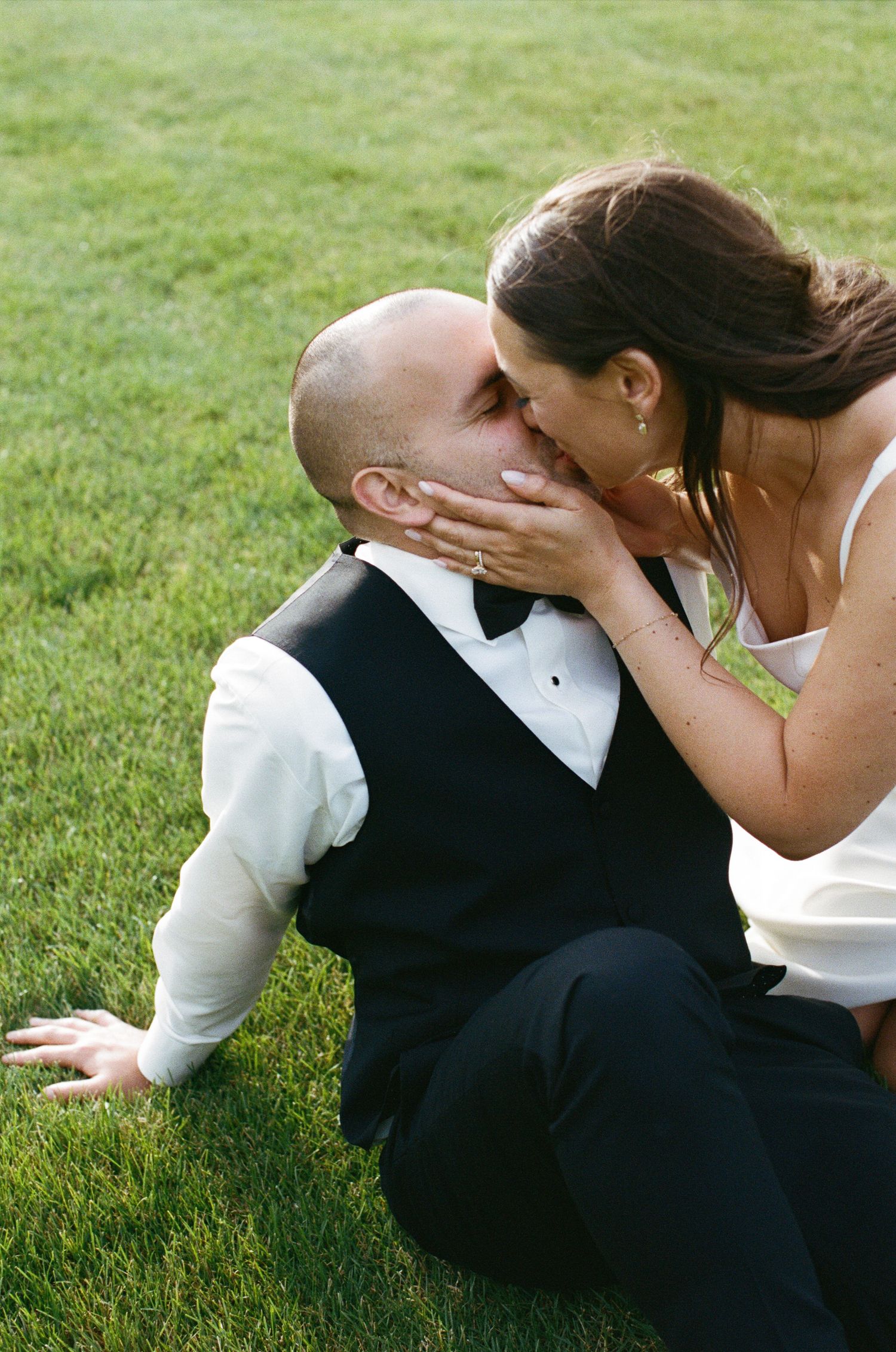 Bride in white gown kisses groom in black vest while sitting on green grass at wedding.