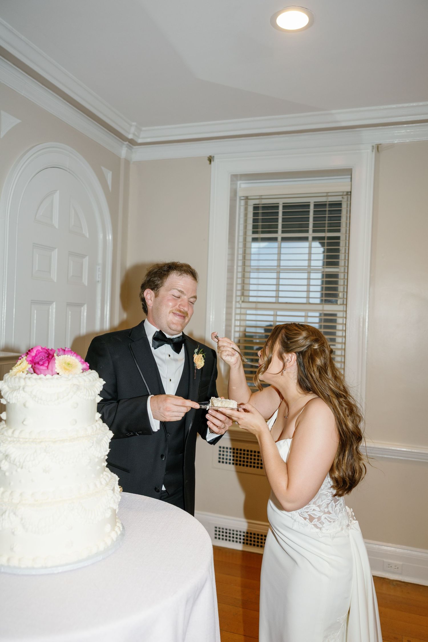 Bride and groom laughing while cutting their white tiered wedding cake decorated with pink flowers.