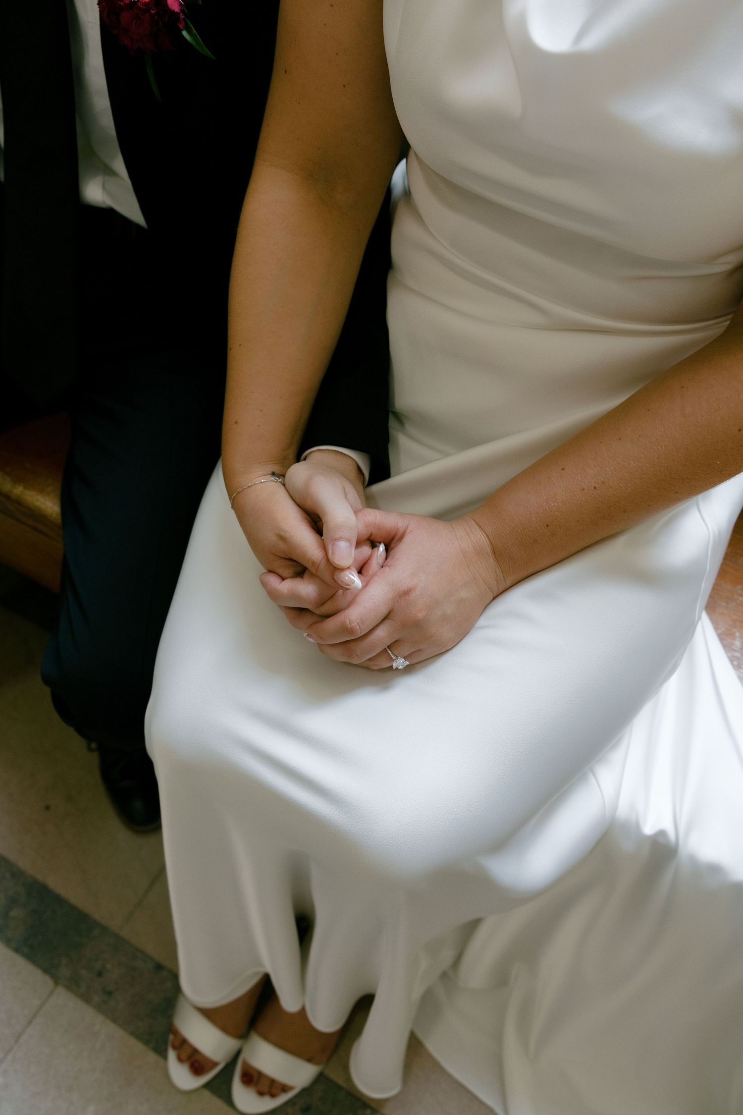 Close-up of bride and groom holding hands, bride wearing engagement ring and white satin gown.