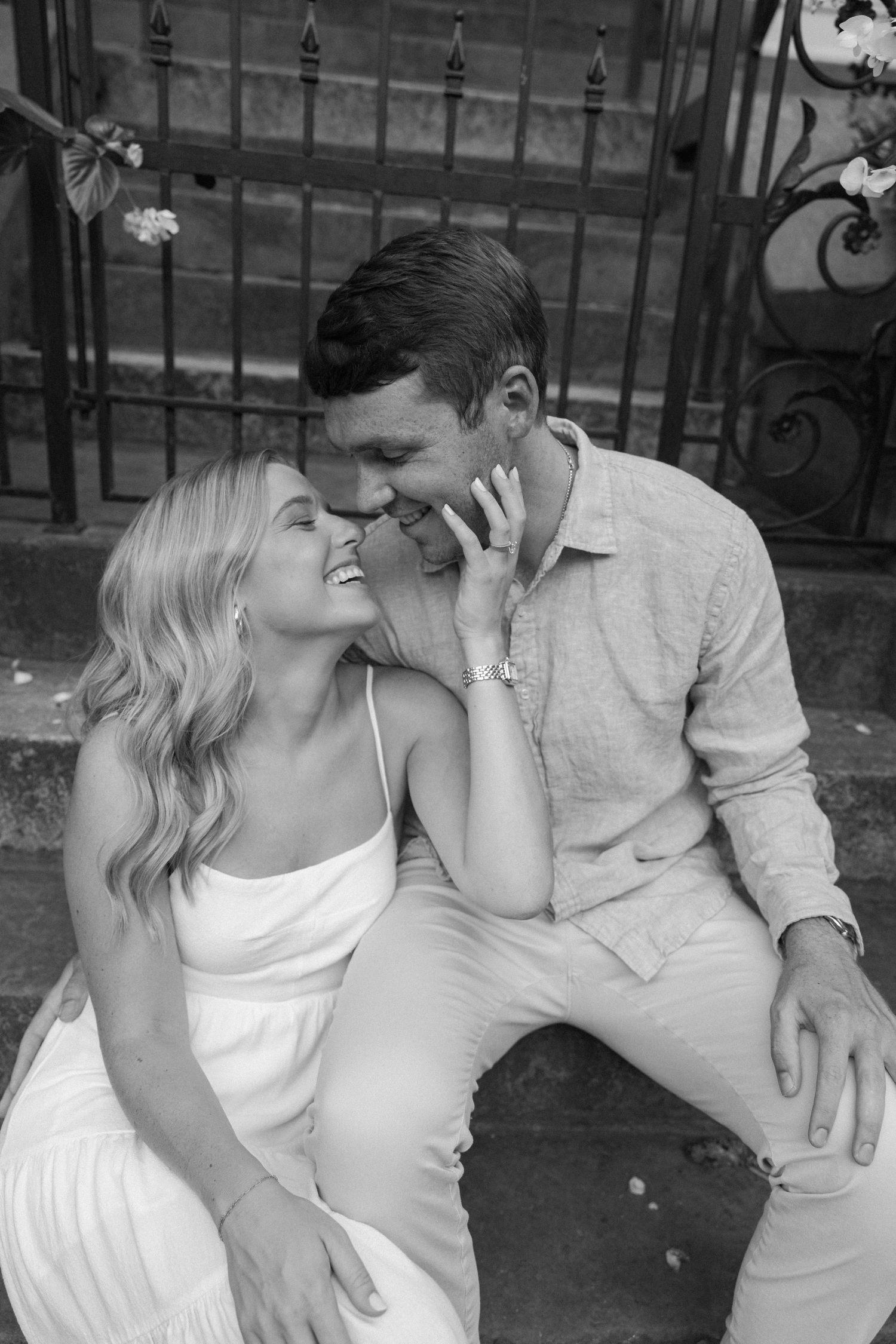 Black and white photo of laughing engaged couple sitting on steps, woman touching mans face tenderly.