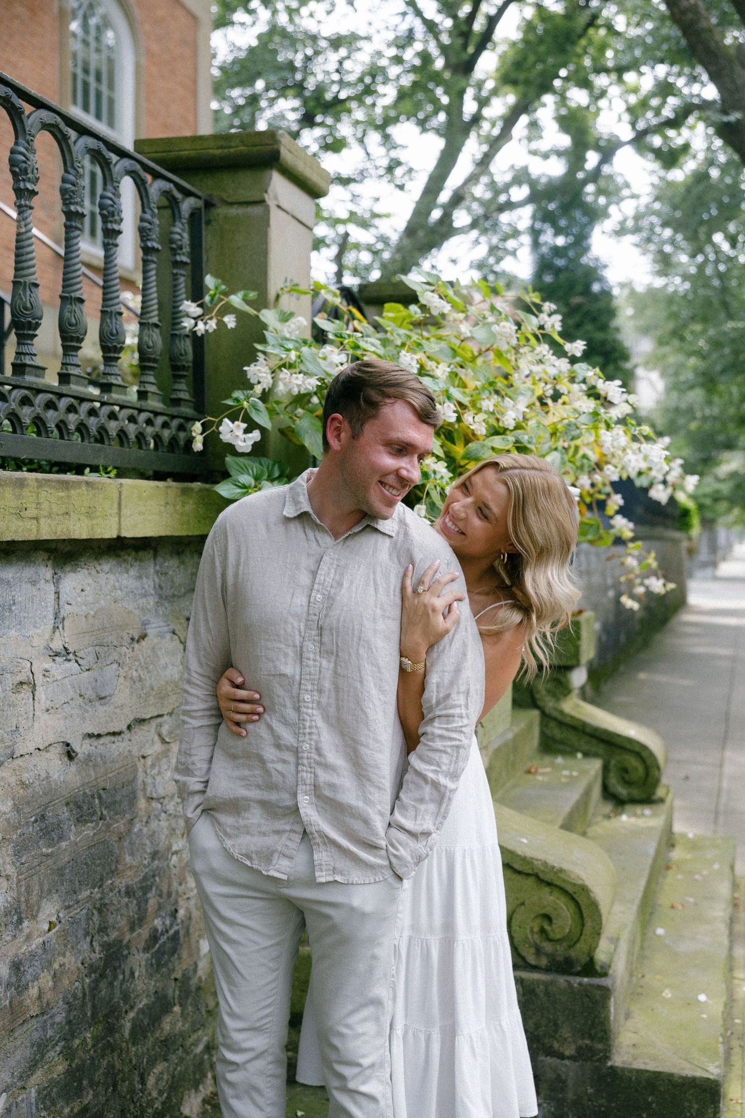 Smiling couple embracing outdoors near stone wall with white flowers, wearing neutral linen outfits.