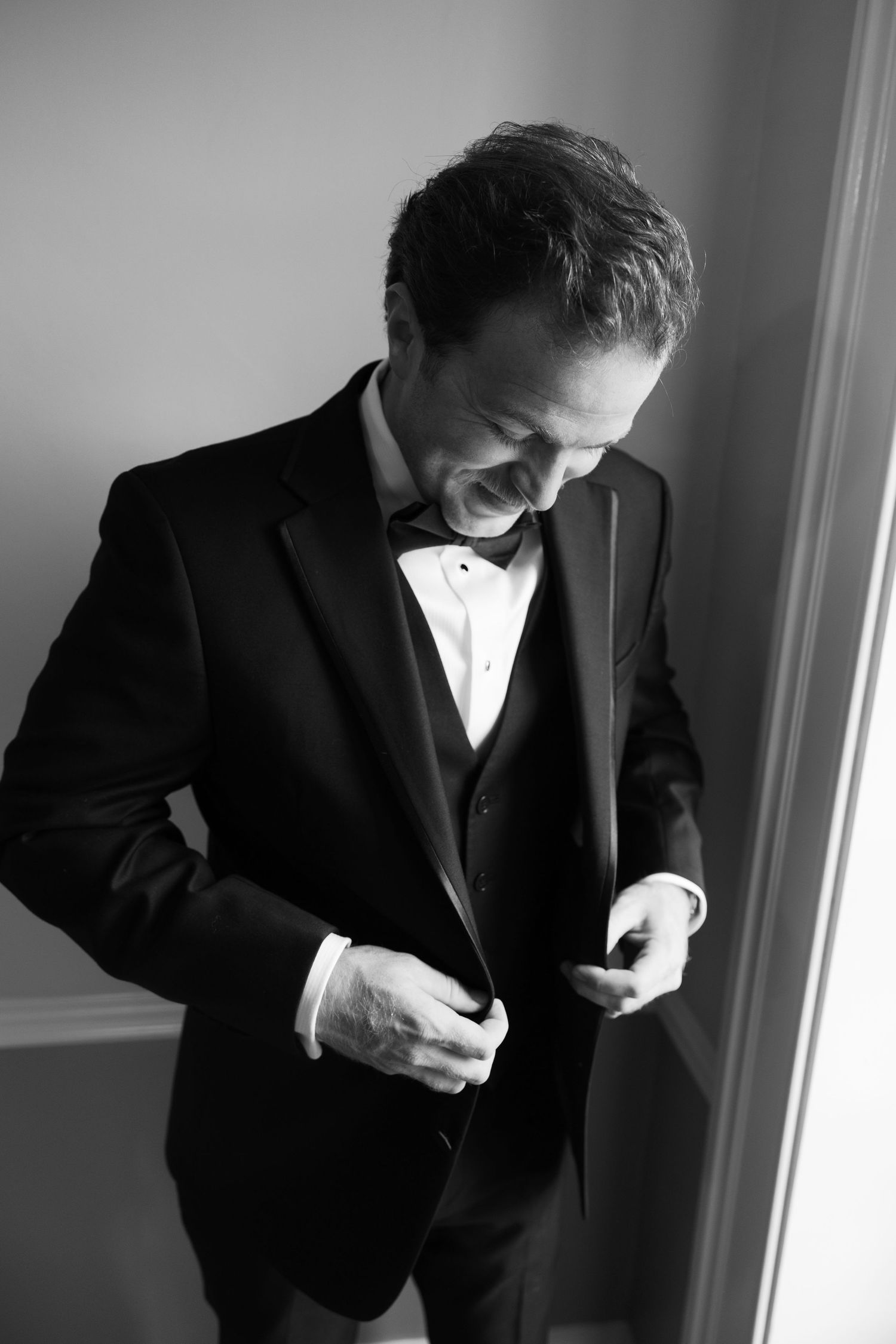 Black and white portrait of a groom buttoning his tuxedo jacket while getting ready for his wedding.
