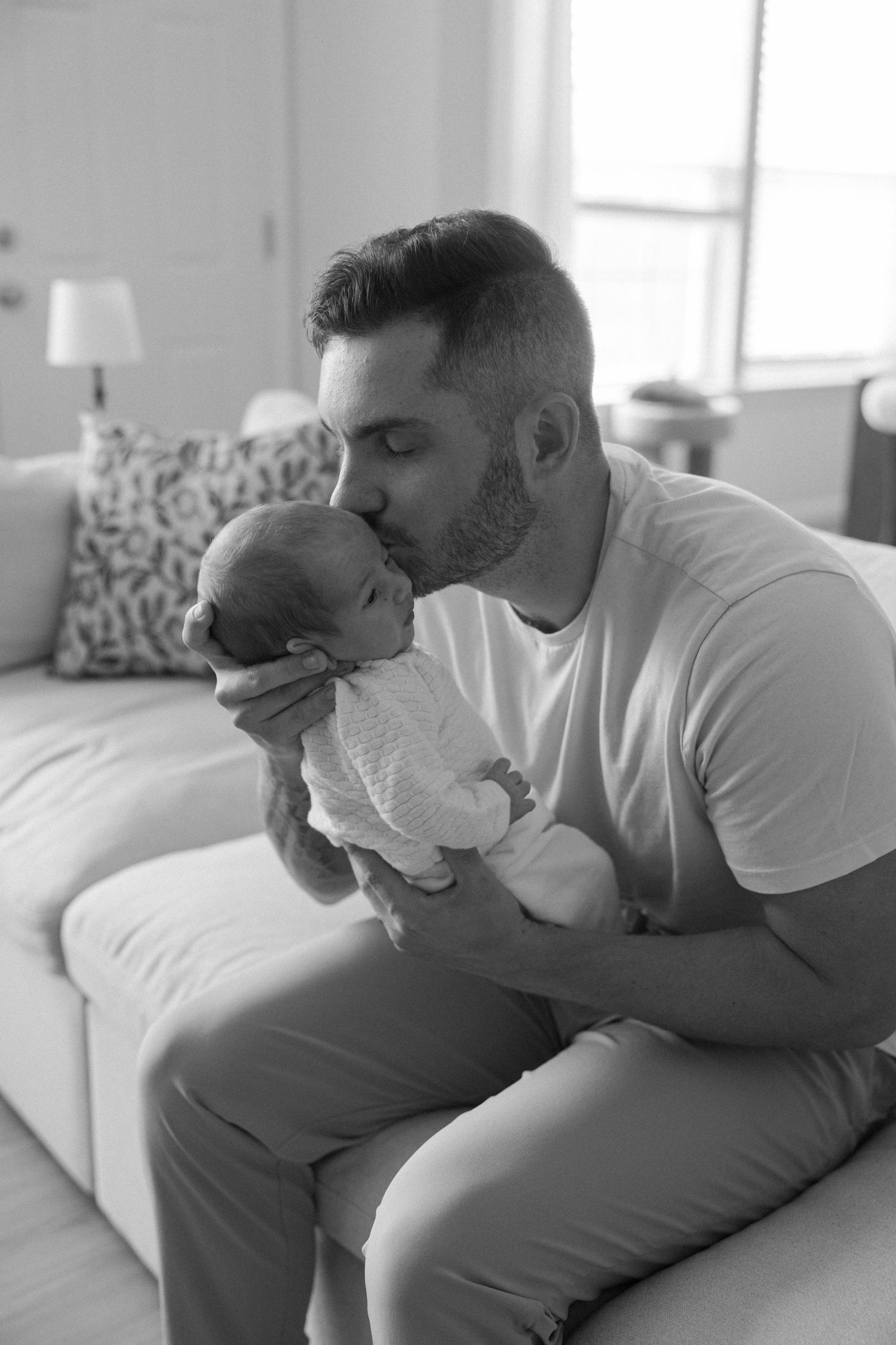 Black and white photo of bearded father kissing newborn baby while seated on couch at home.