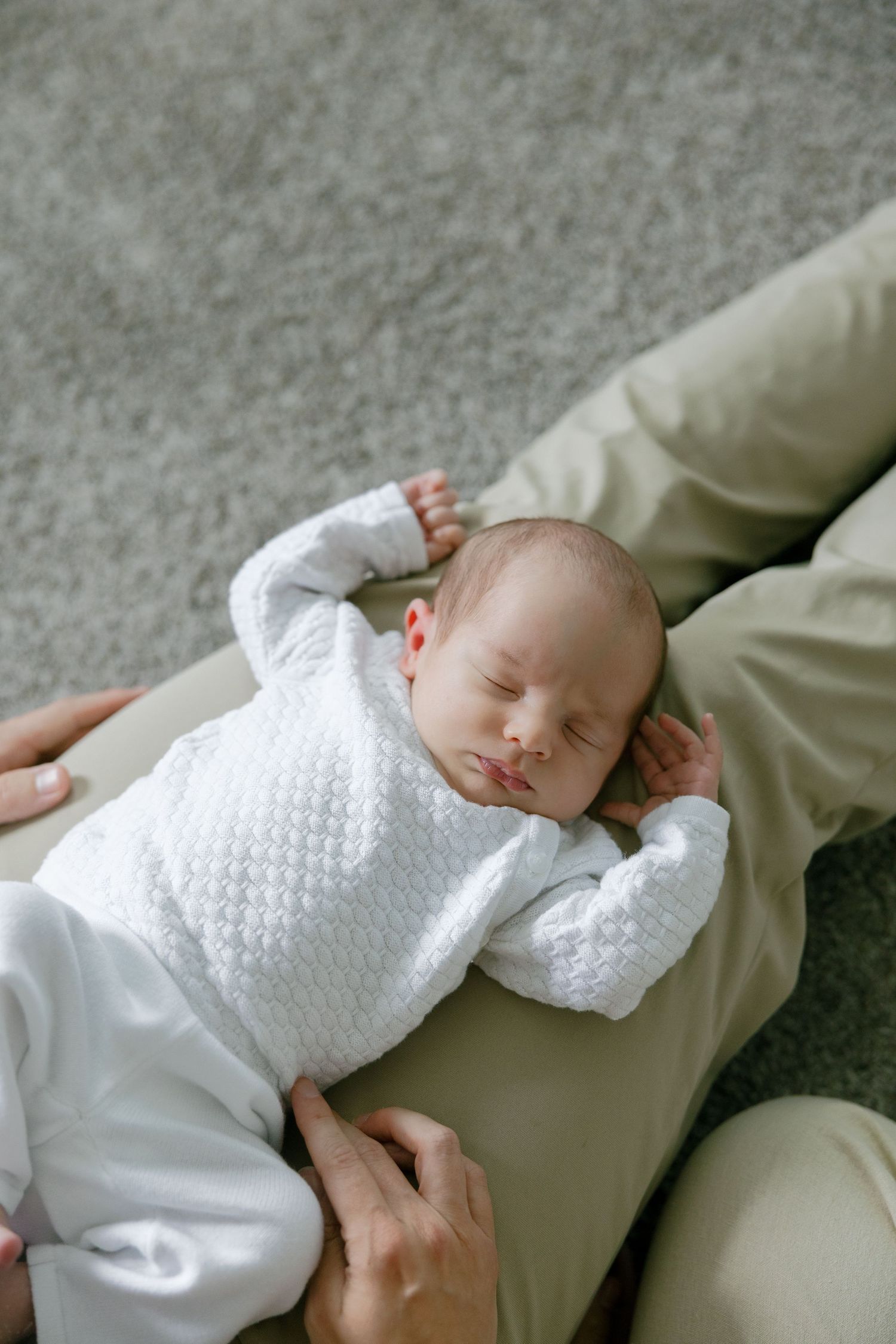 Sleeping newborn baby in white knit outfit resting peacefully on parent's lap on carpet floor.