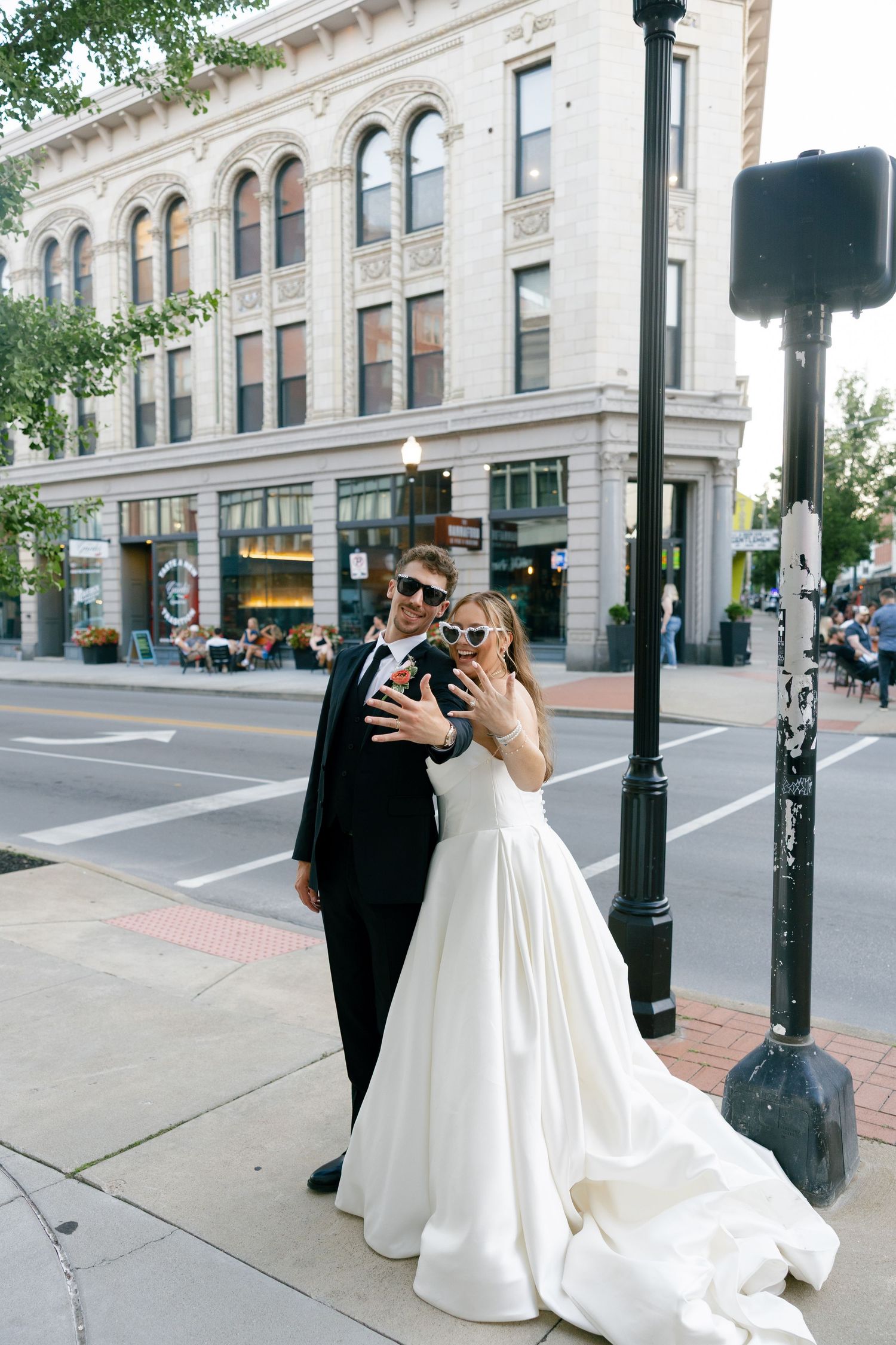 Bride and groom wear sunglasses and pose playfully on a city sidewalk with a historic building in the background.