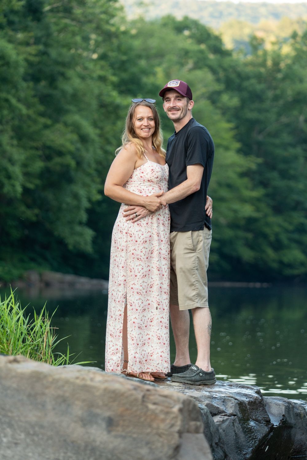 Couple poses on rocky shoreline beside tranquil lake during outdoor photoshoot.