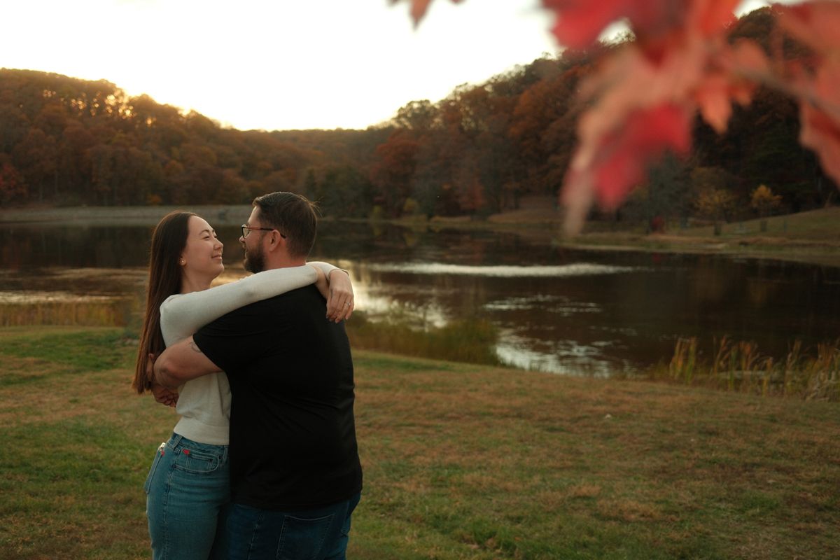 Couple embracing at sunset with autumn trees and lake in background.