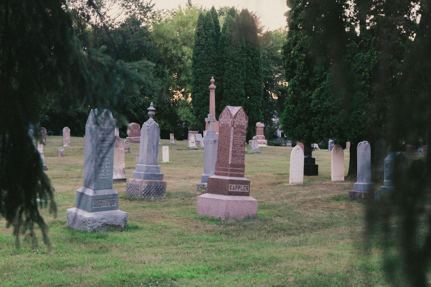 Historic cemetery with weathered gravestones and monuments surrounded by lush green trees at dusk.