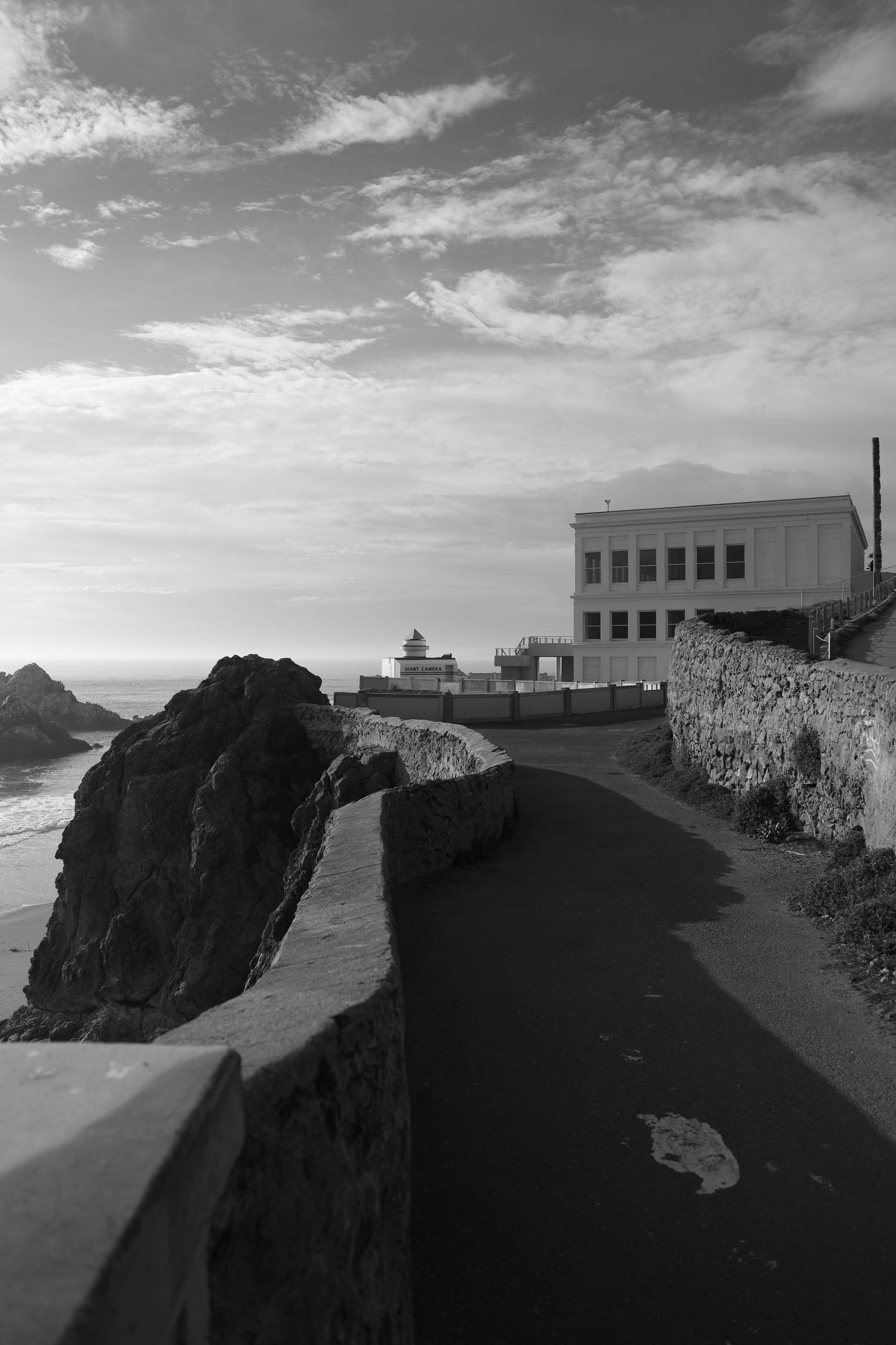 Black and white coastal path leading to a historic building at the edge of the ocean at sunset.