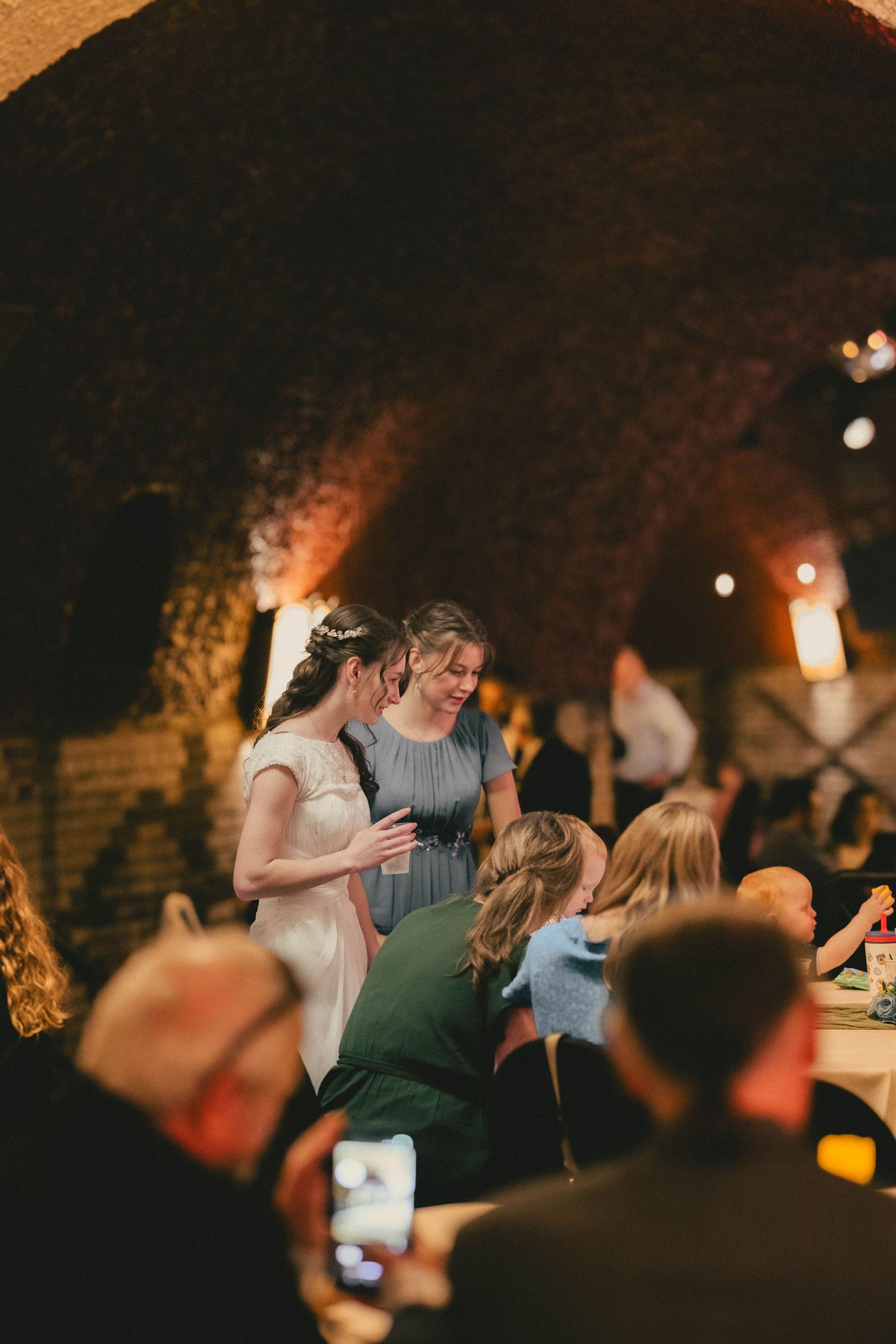 Two women share a warm moment at an elegant wedding reception in a rustic brick vaulted cellar venue.