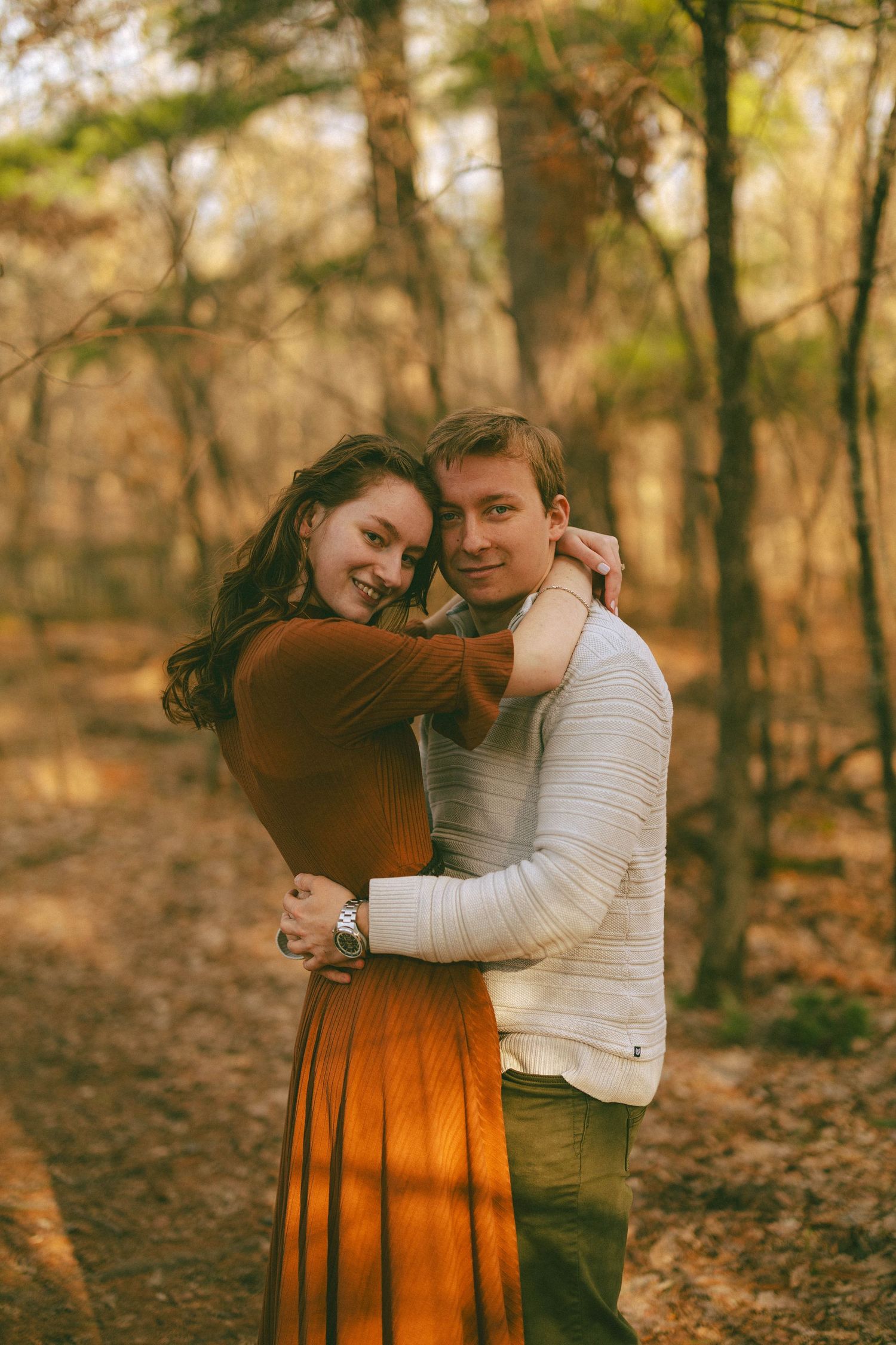 Smiling couple embracing in a warm autumn forest, woman in rust dress wrapping arms around mans neck.