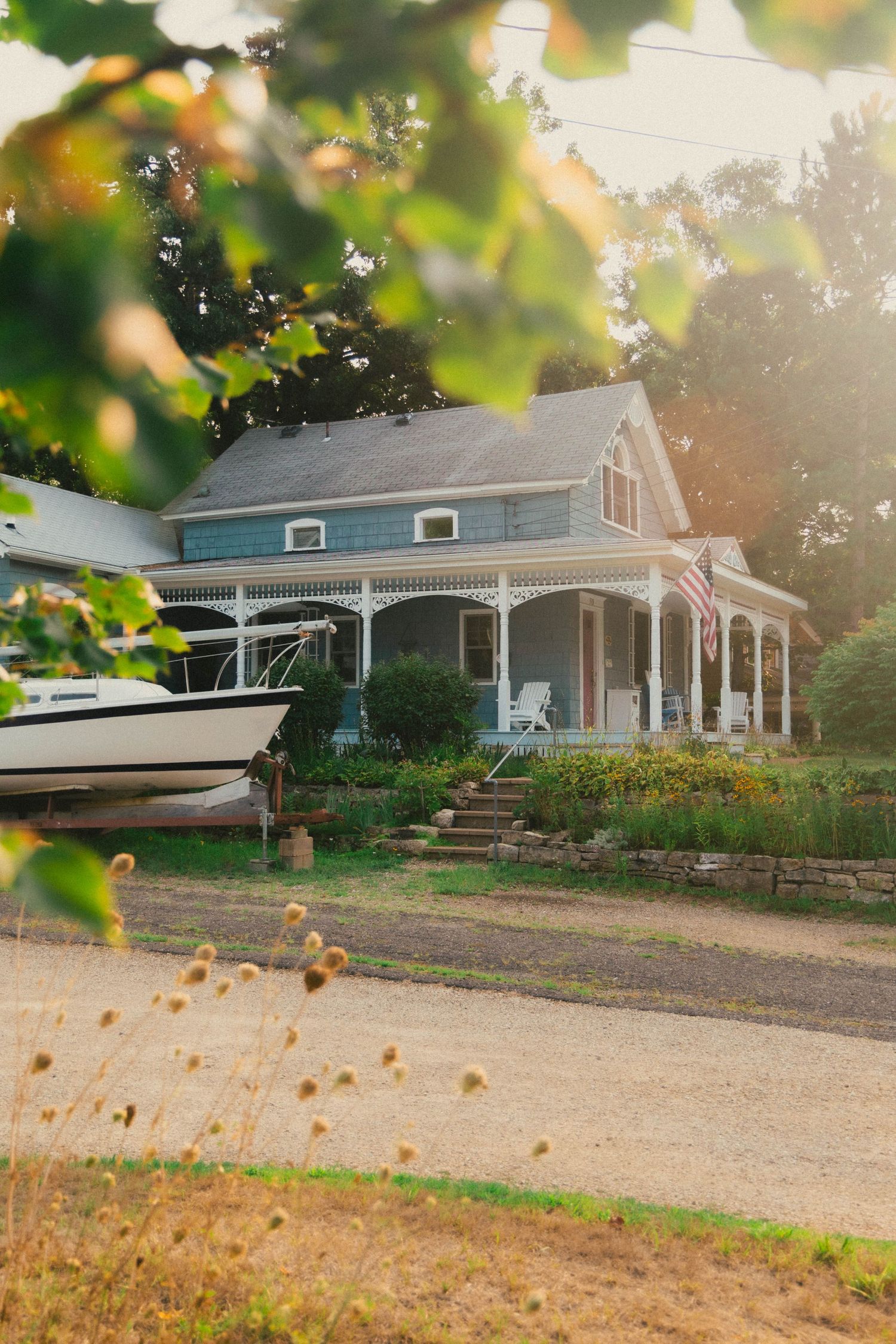 Charming Victorian farmhouse with wrap-around porch, American flag, and boat in driveway, bathed in warm golden sunset light.