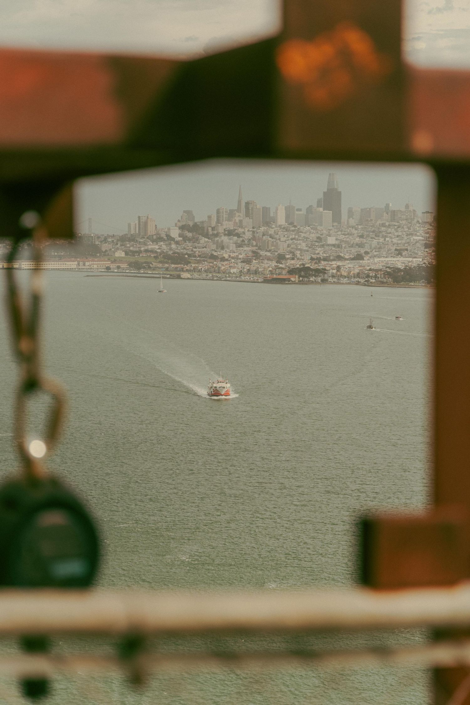 A boat leaving a white wake across San Francisco Bay viewed through a metal bridge frame with city skyline beyond.