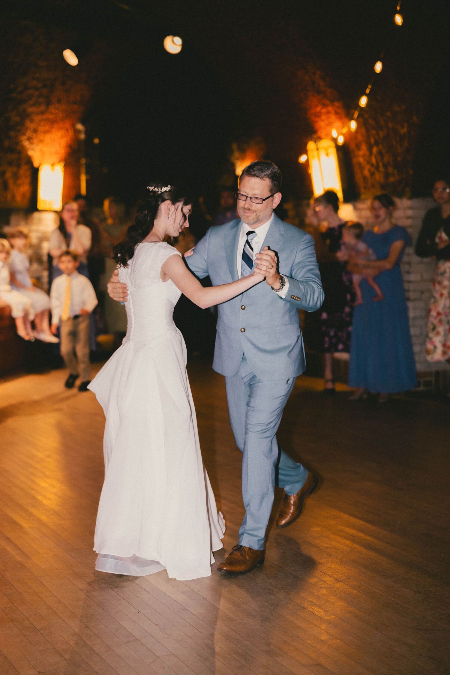 Bride and groom share first dance on wooden floor at elegant wedding reception with warm ambient lighting.
