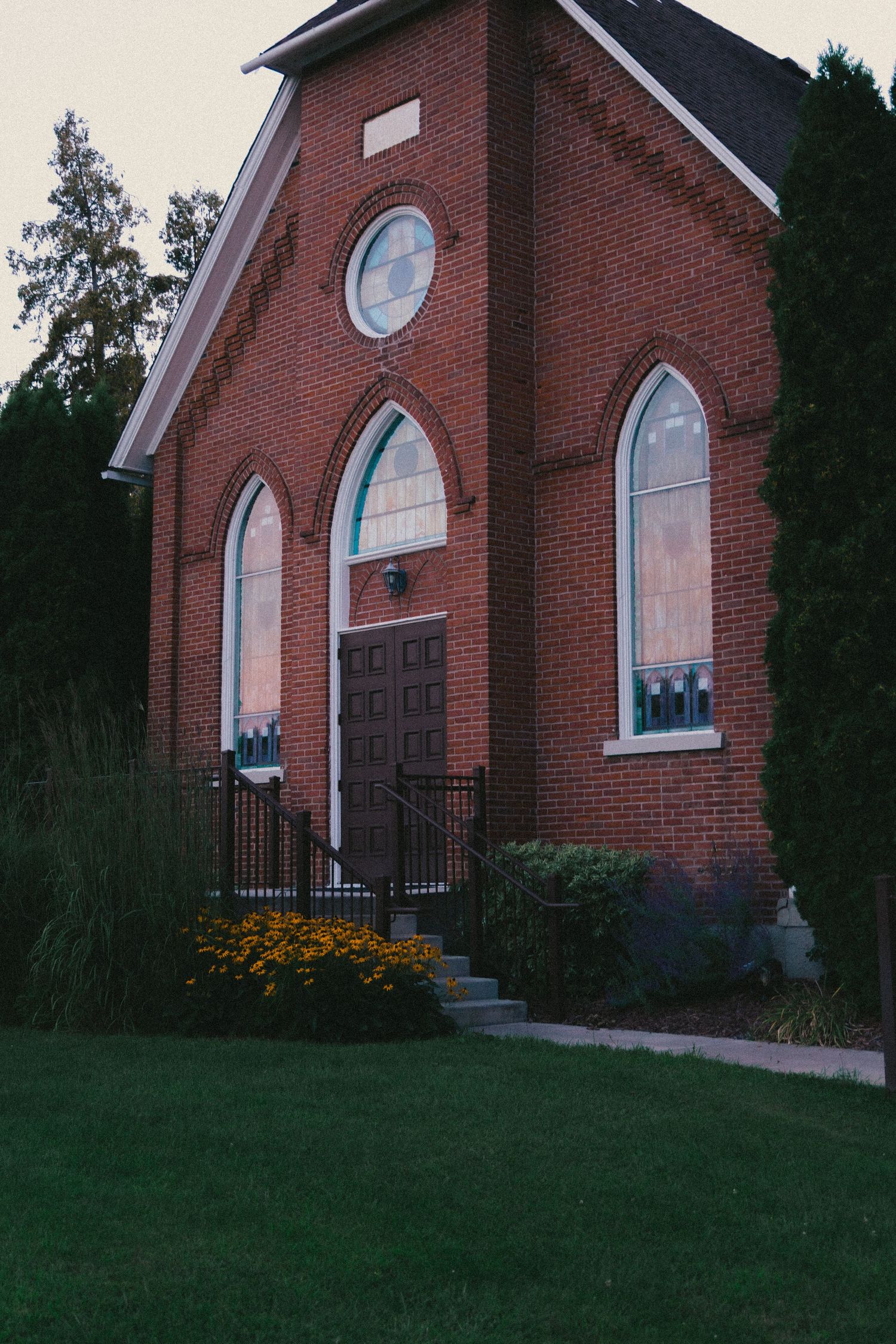 Red brick church with arched windows, circular window, wooden door, iron railings, yellow flowers, and lush green lawn at dusk.