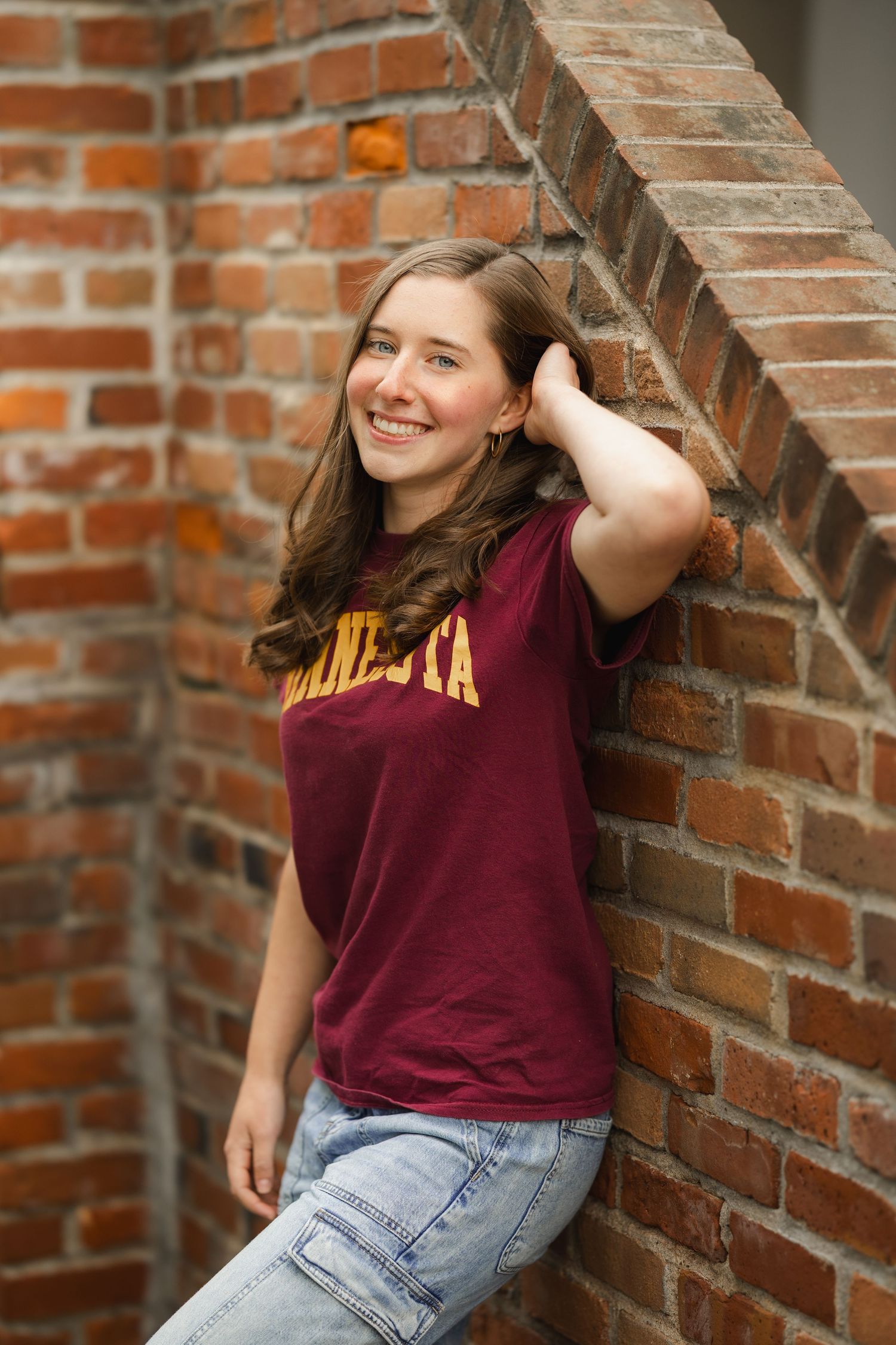 Smiling teenage girl in a maroon Minnesota t-shirt leaning against a brick wall in a casual senior portrait.