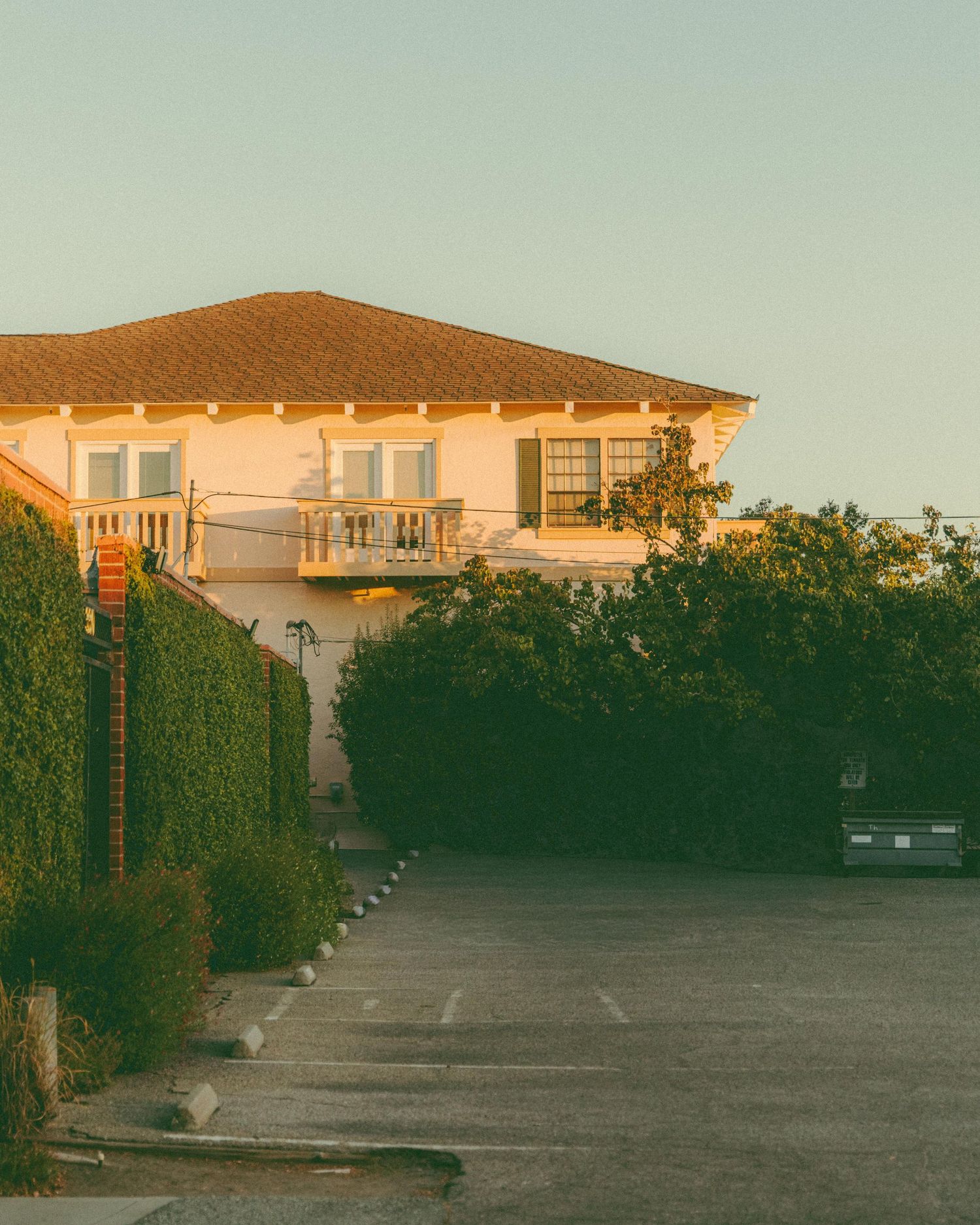 A two-story white house with a balcony bathed in warm golden sunset light, surrounded by lush green hedges and an empty parking lot.
