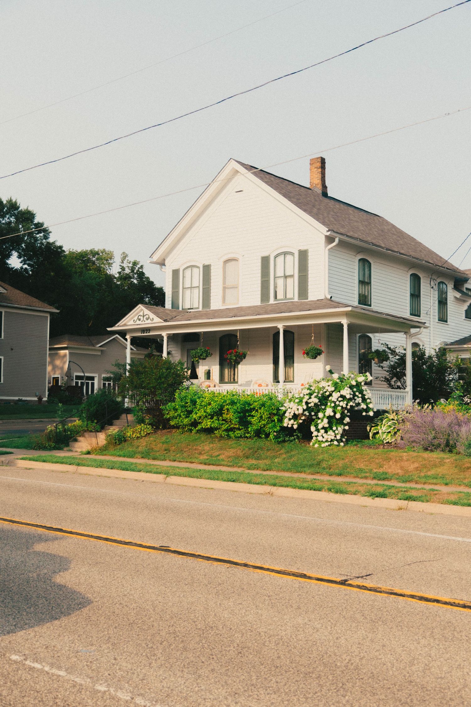 White two-story farmhouse with green shutters, wrap-around porch, and blooming gardens along a quiet small-town street.