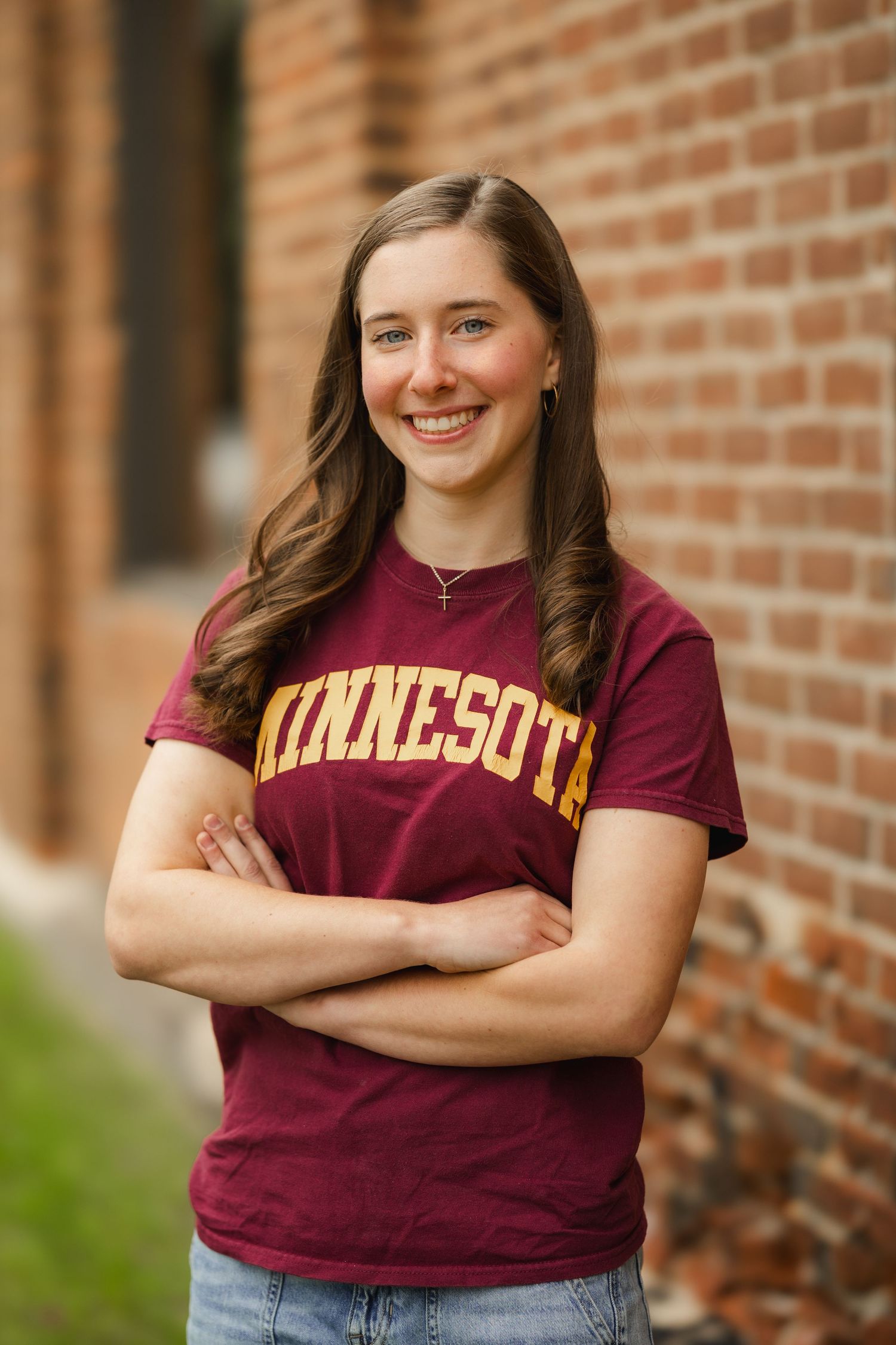 Smiling young woman with crossed arms wearing a maroon Minnesota Gophers t-shirt posed against a brick wall.
