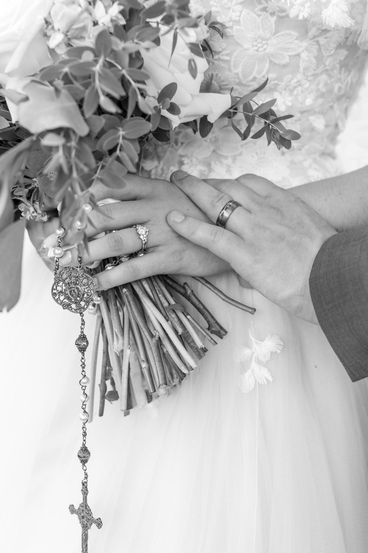 Black and white close-up of bride and groom hands with wedding rings holding floral bouquet with rosary.