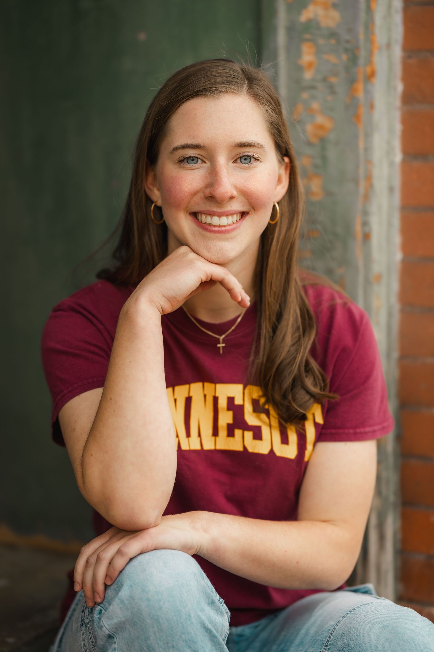 Smiling young woman in a maroon Minnesota t-shirt sitting casually with hand on chin, wearing gold hoop earrings and a cross necklace.