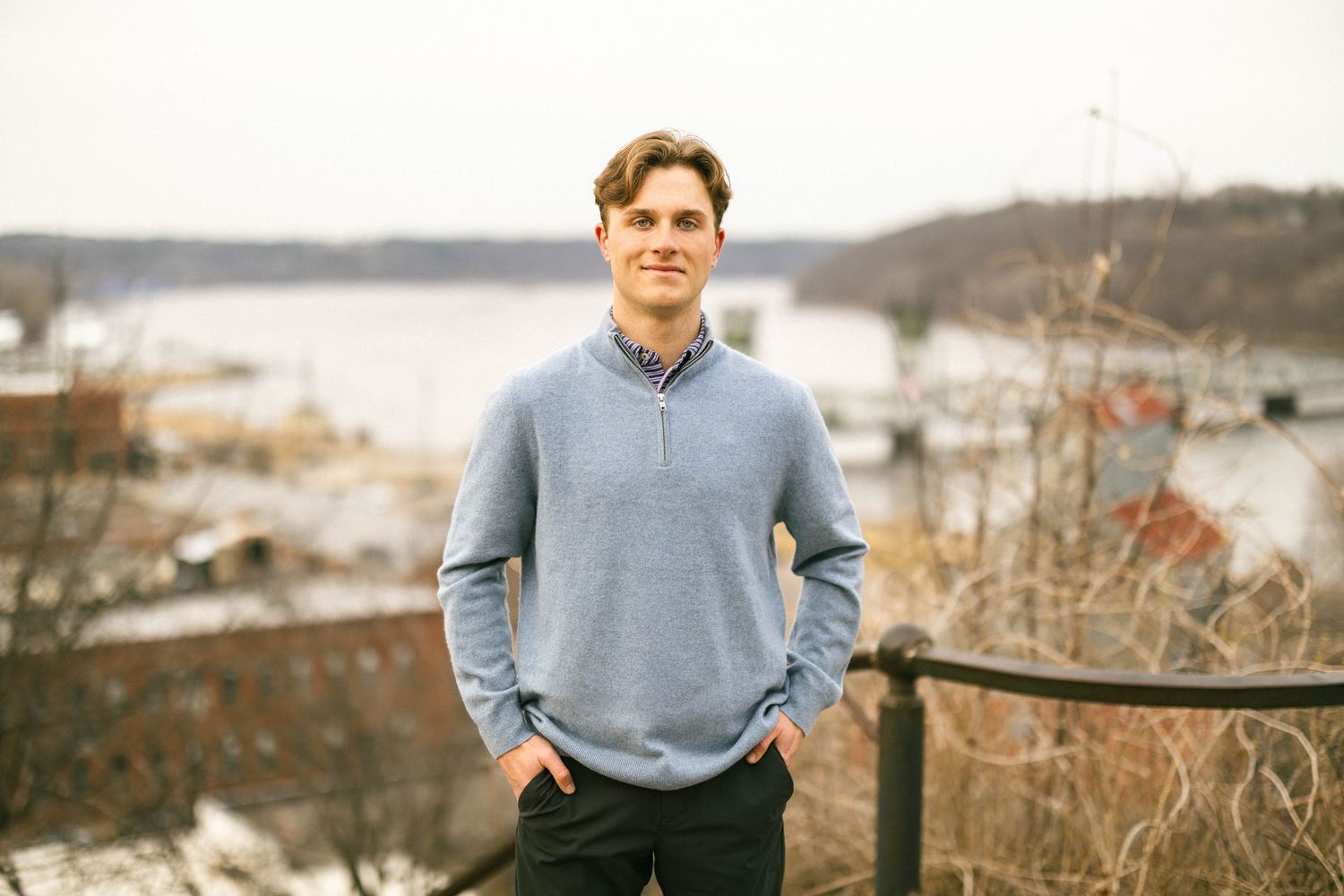 Young man in gray quarter-zip pullover standing on overlook with river view.