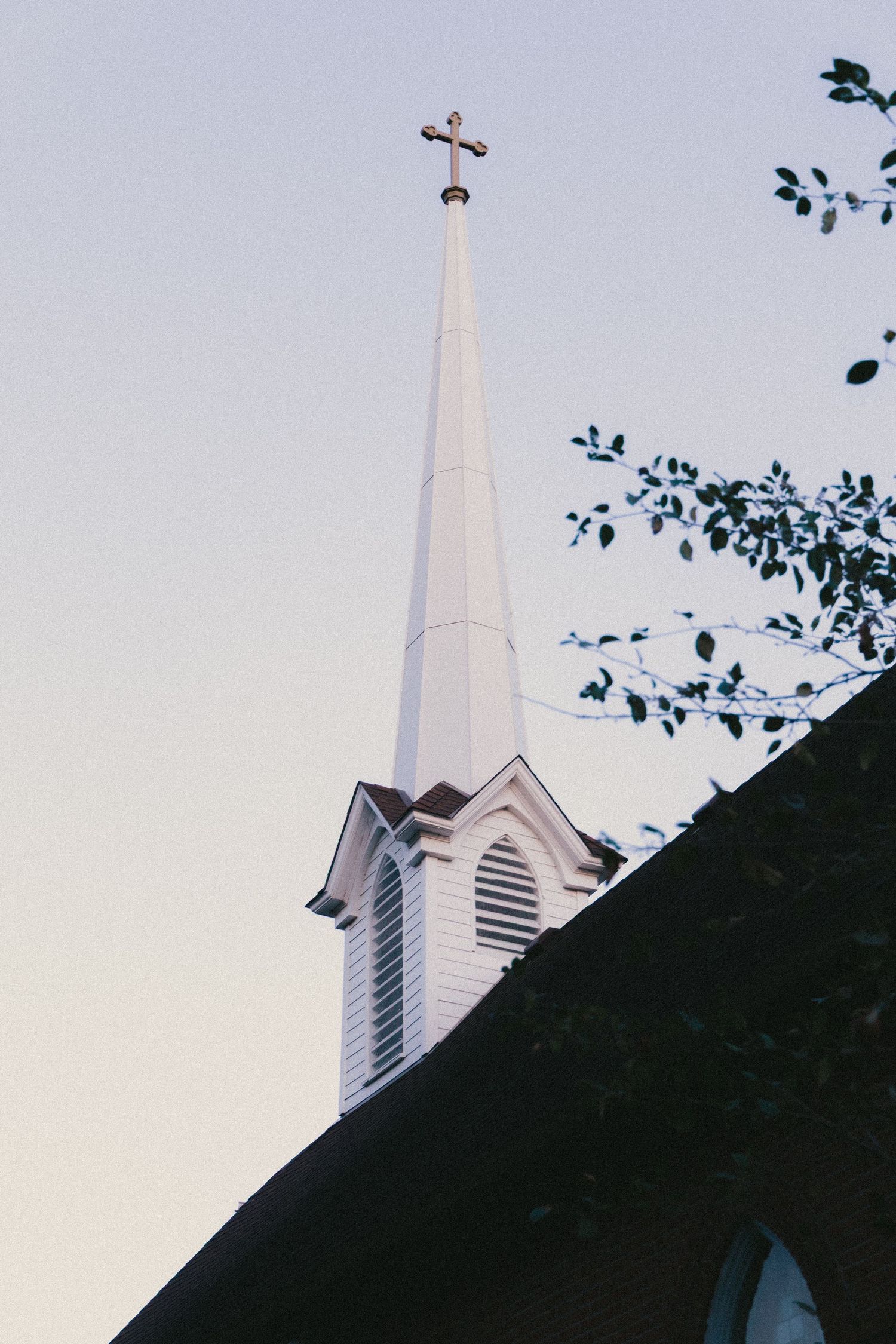 White church steeple with a cross on top against a clear sky, framed by tree branches.