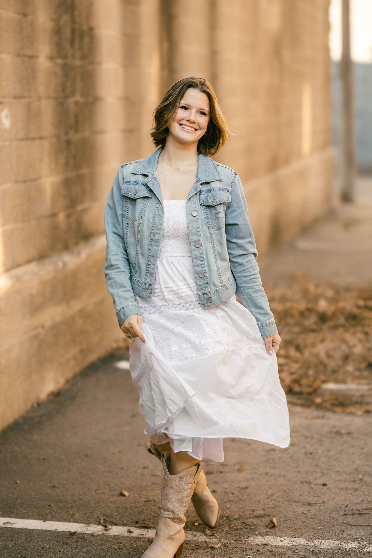 Woman in white dress, denim jacket, and tan boots smiling while walking near a stone wall outdoors.