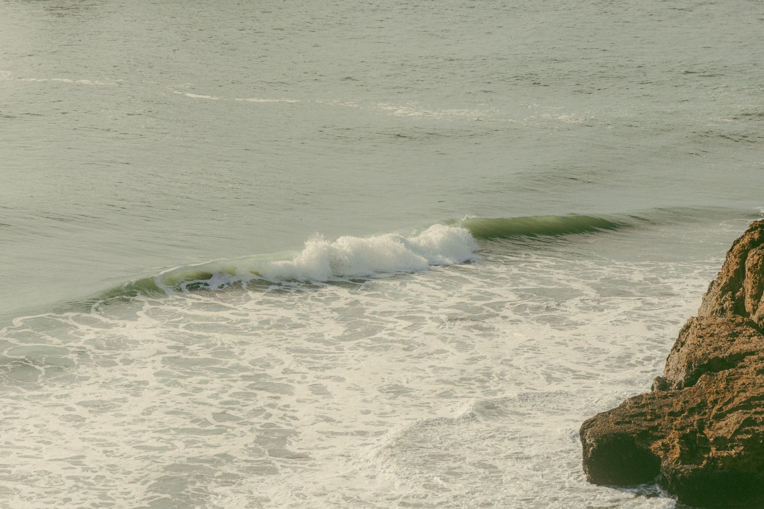 Ocean waves crashing against rocky coastline with white sea foam in muted green tones at dusk.