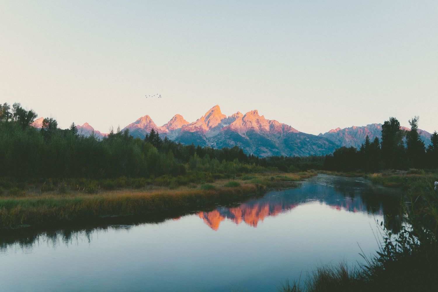 Serene mountain landscape at sunset with pink peaks reflecting in calm river waters surrounded by lush green forest.