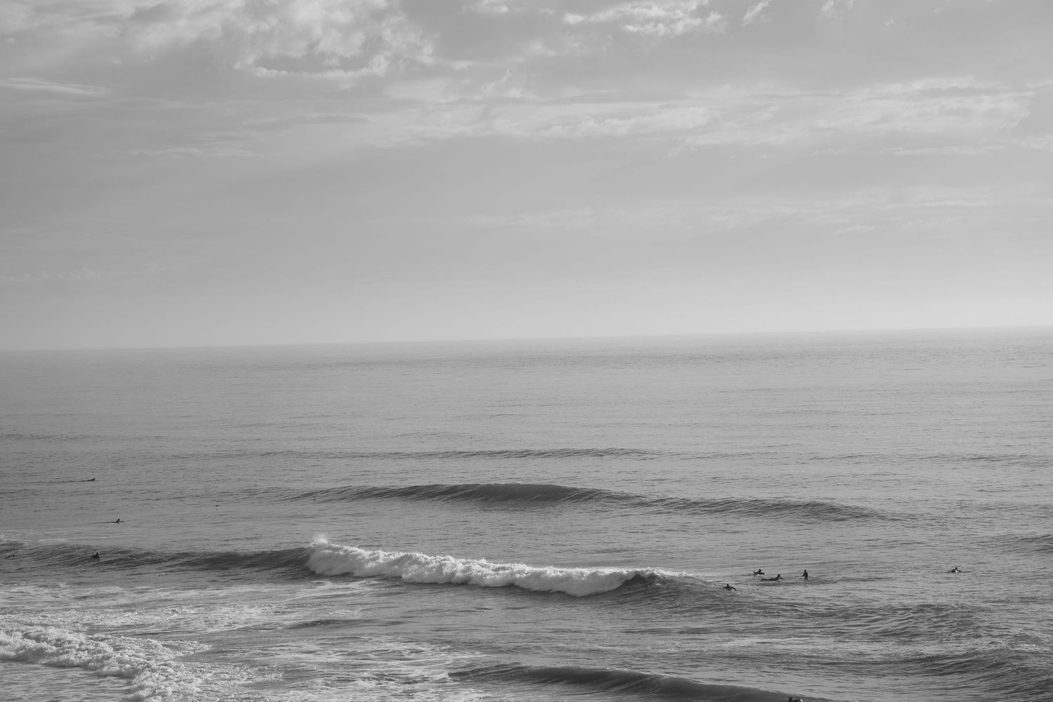 Black and white ocean seascape with gentle waves and surfers in the distance under a cloudy sky.