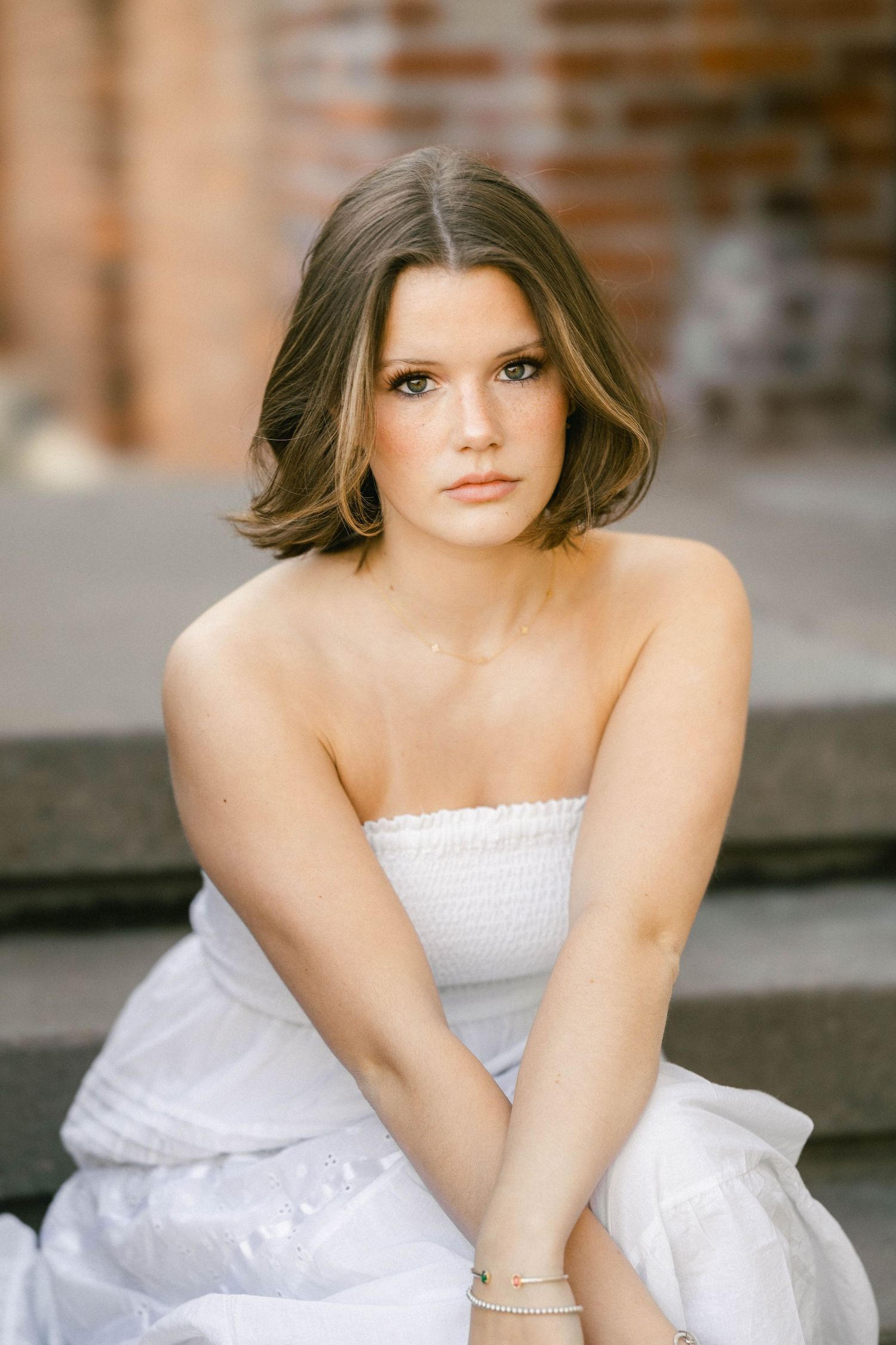 Young woman in white strapless dress with short brown hair sitting on steps near brick wall.