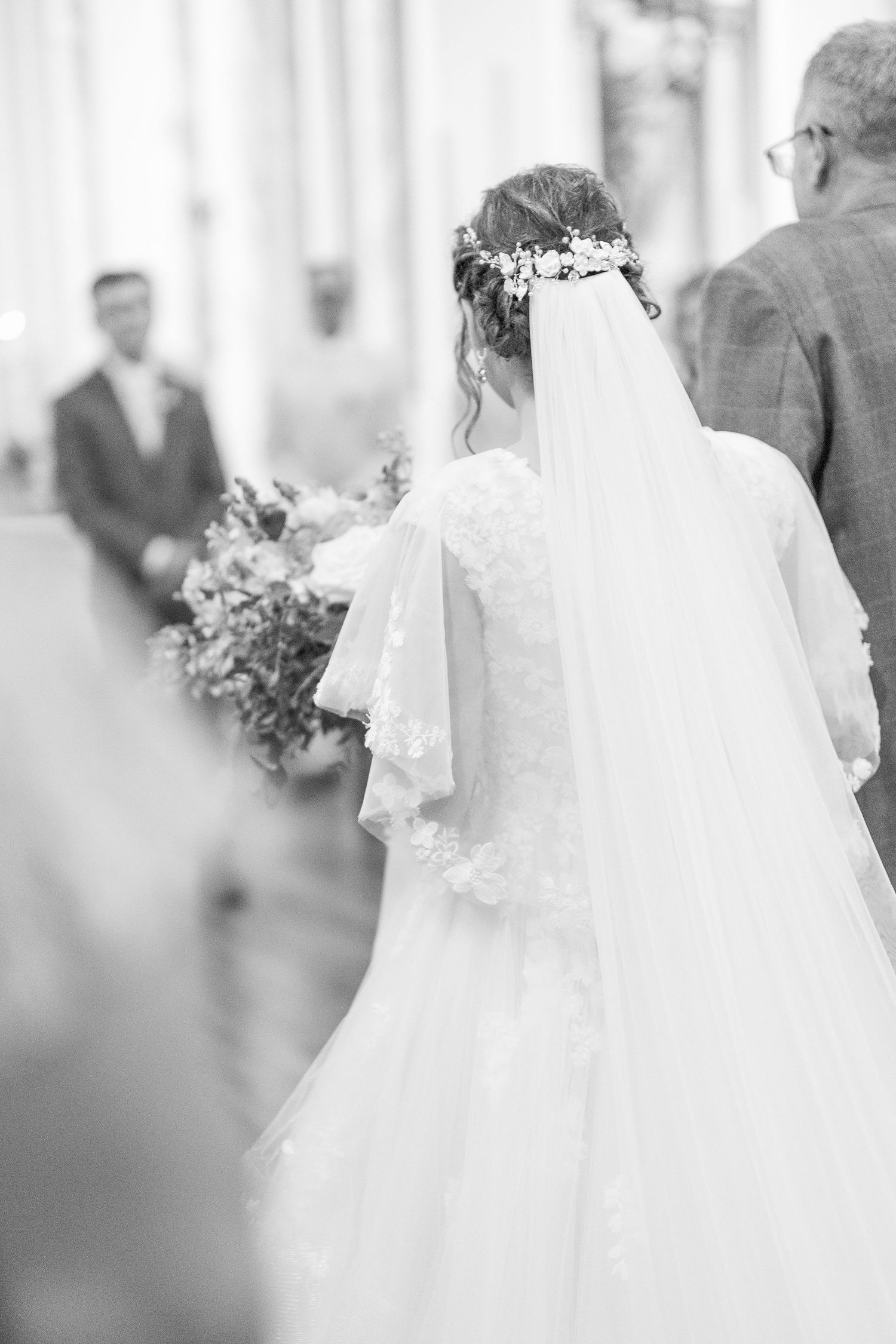 Elegant bride in a lace gown and floral crown walking down the aisle in black and white.