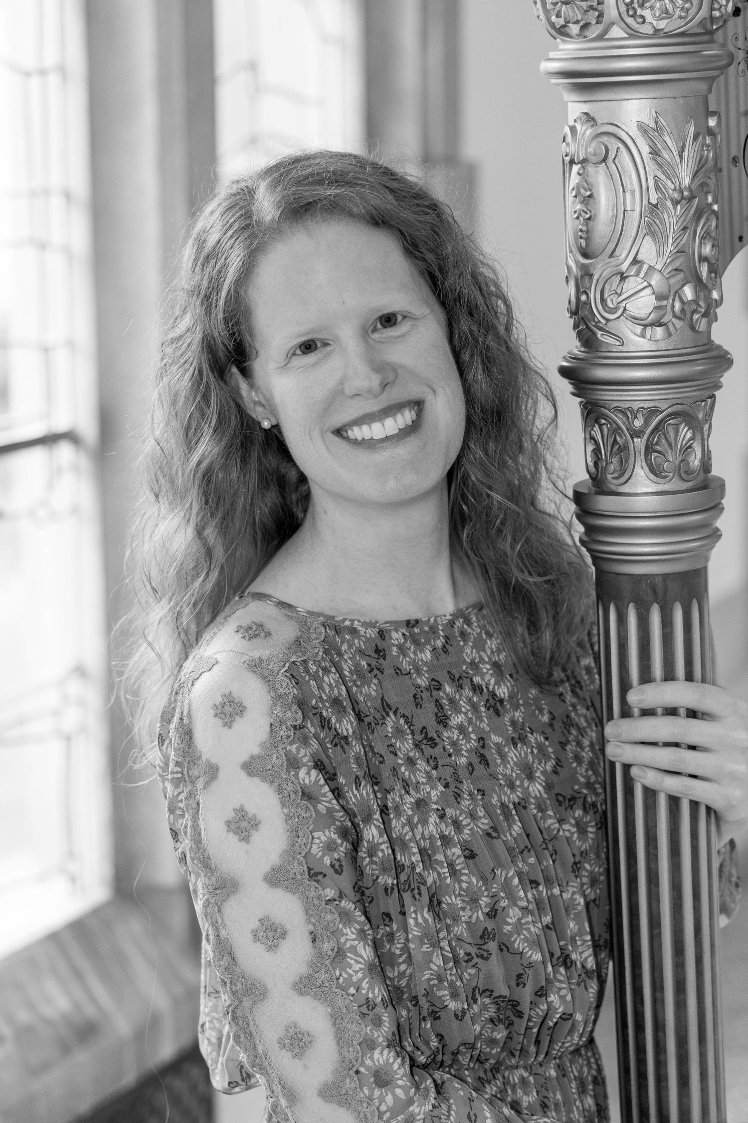 Black and white portrait of a smiling woman with curly hair posing beside an ornate decorative harp column.