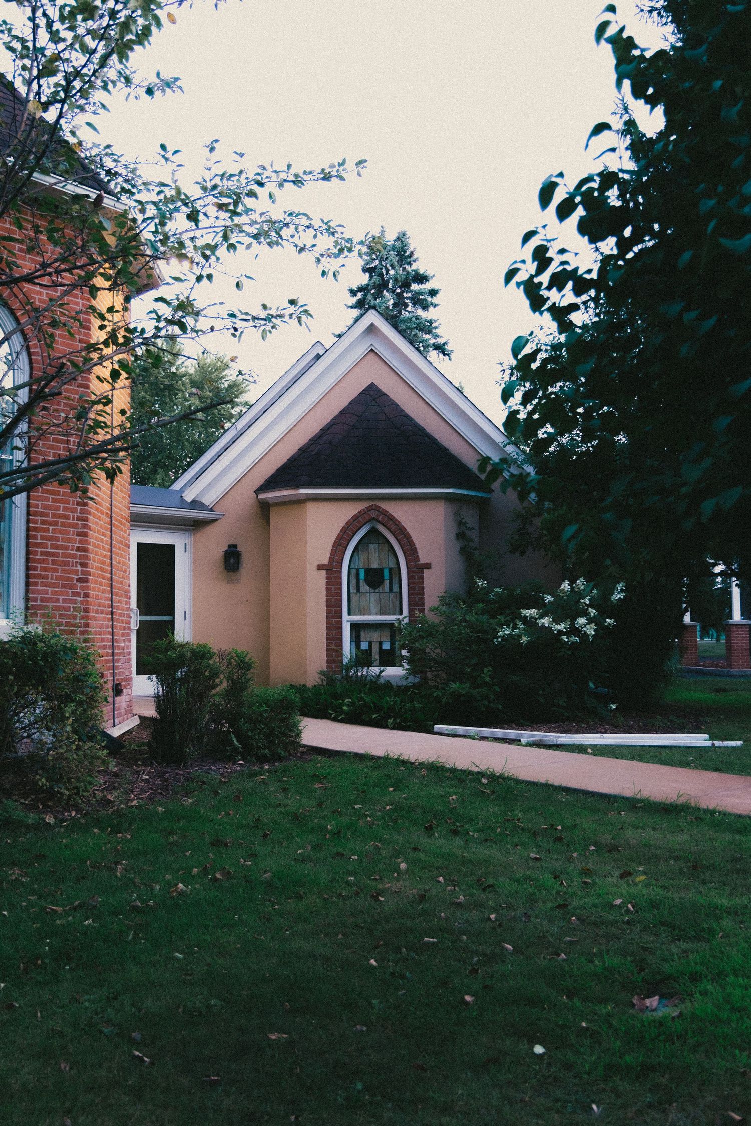 A charming yellow stucco church entrance with Gothic arch window, brick facade, and lush green trees surrounding the pathway.