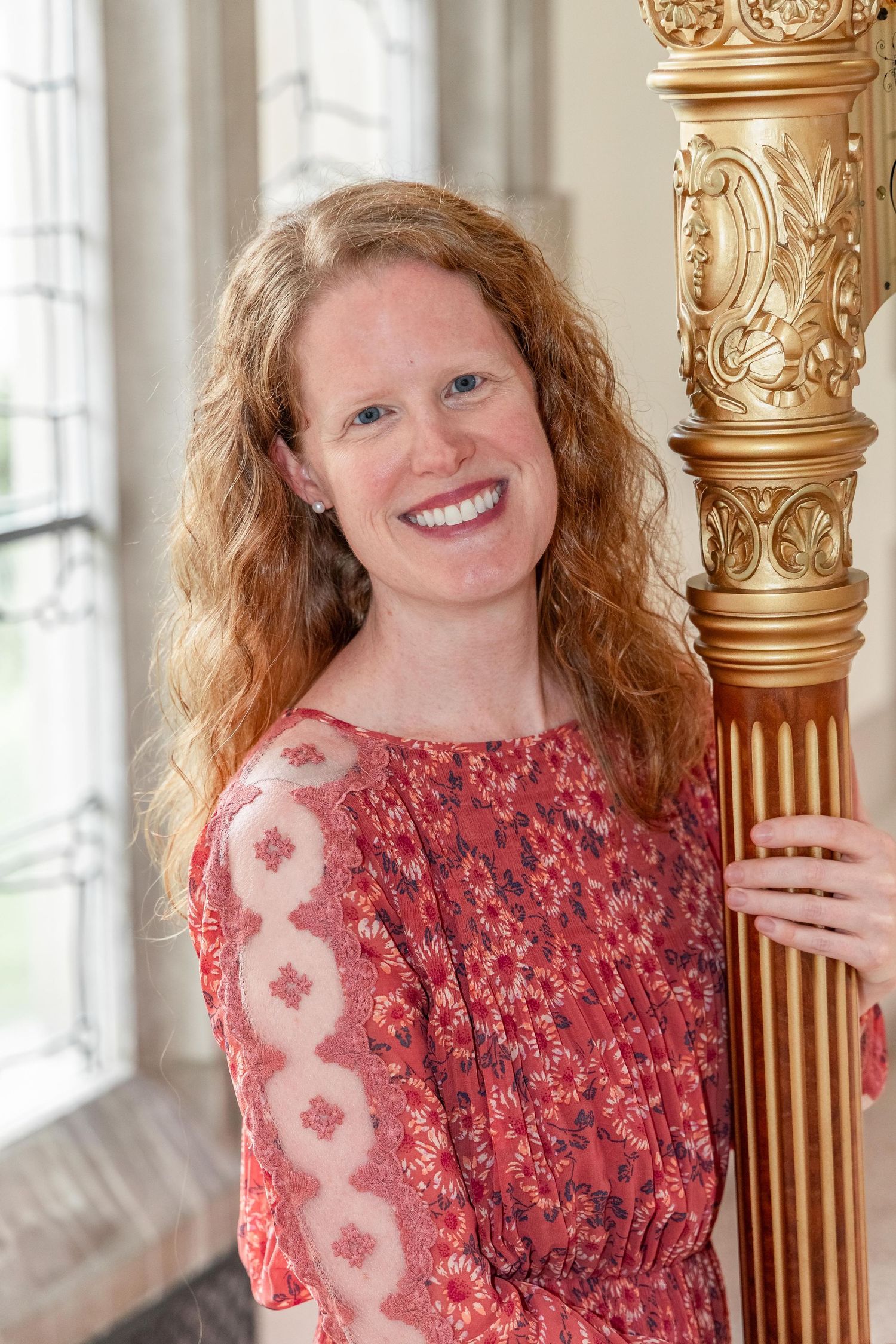 A smiling woman with curly red hair in a floral dress poses beside a ornate gold harp in a bright room.