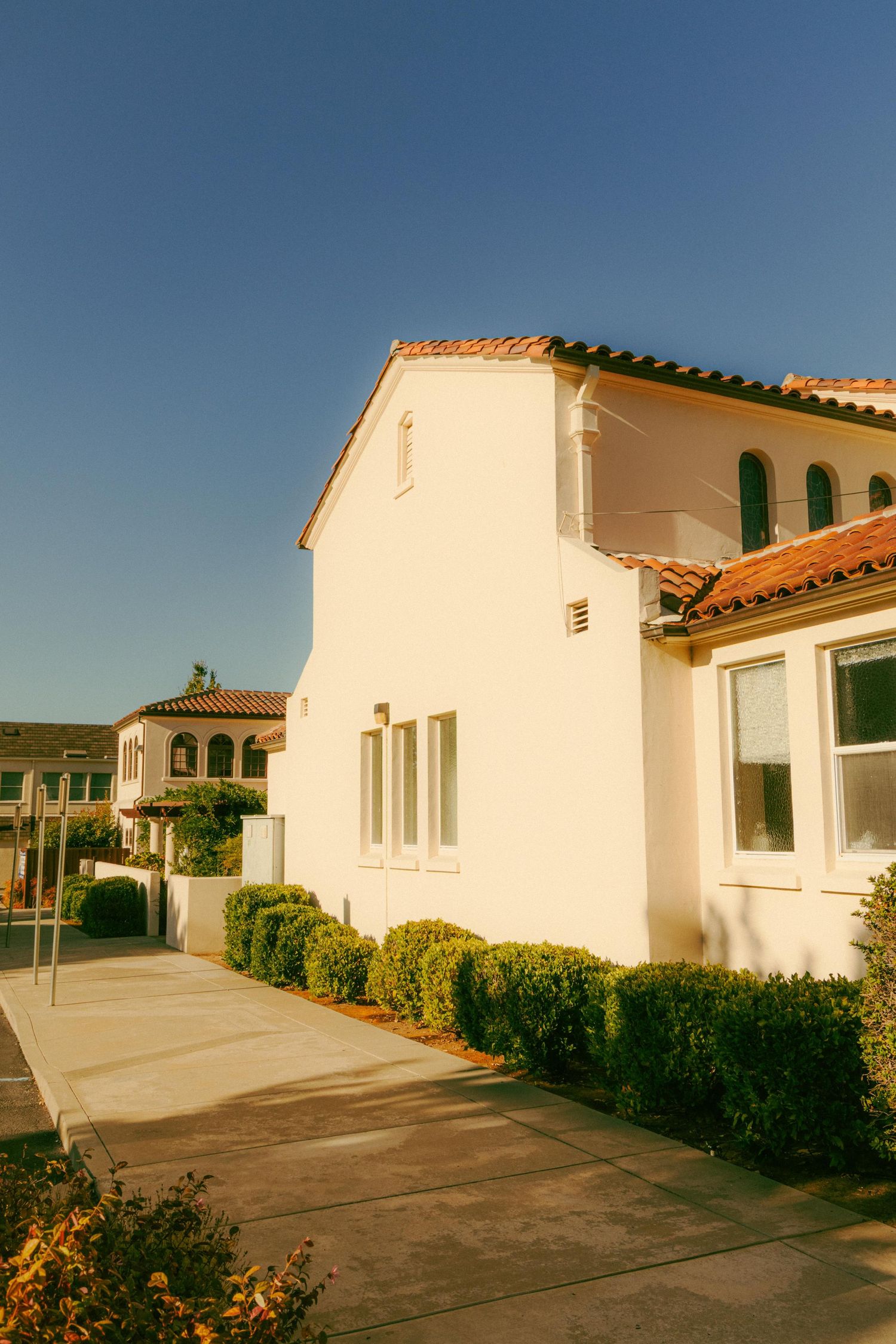 White Spanish Colonial Revival style home with red tile roof and manicured hedges under a clear blue sky.