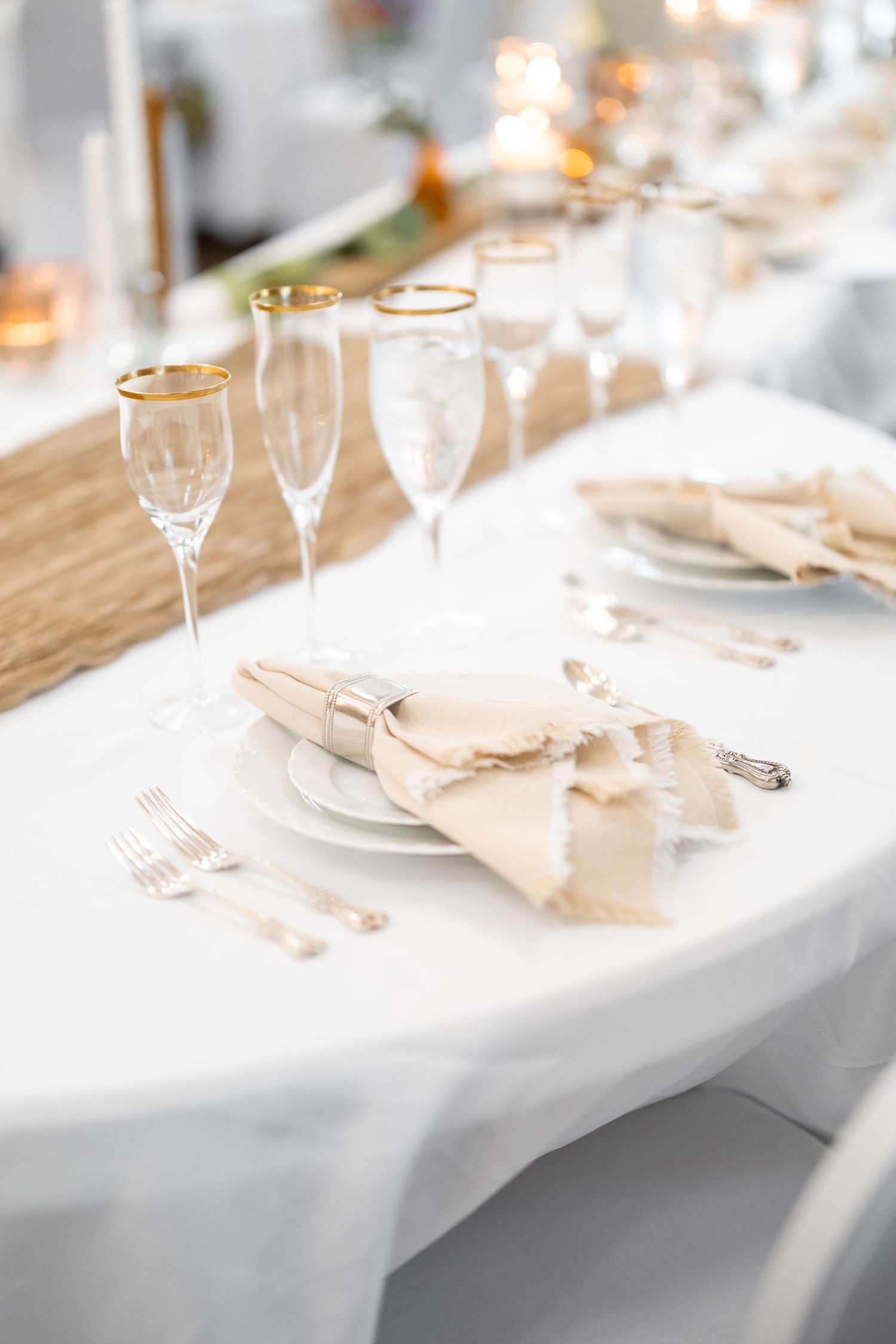 Elegant wedding place setting with champagne linen napkin, gold-rimmed glassware, and silver cutlery on white tablecloth.