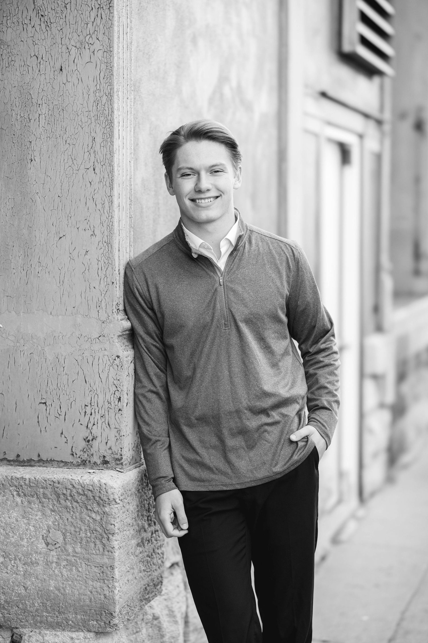 Young man smiling in black and white photo, wearing a quarter-zip pullover, leaning against a stone building wall.