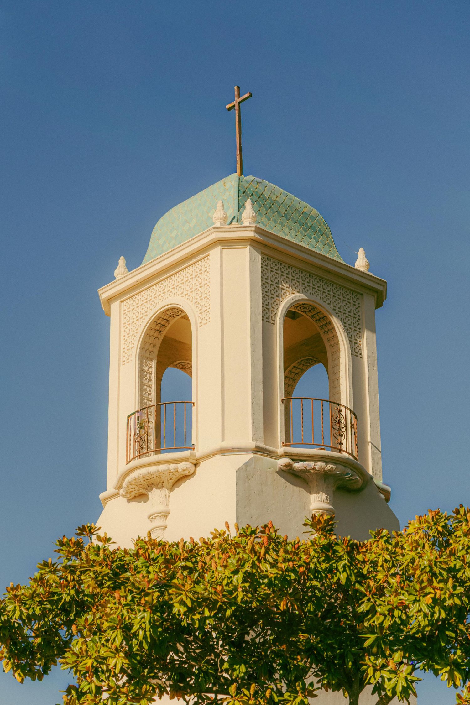 Ornate octagonal church bell tower with green dome and cross against a clear blue sky framed by trees.