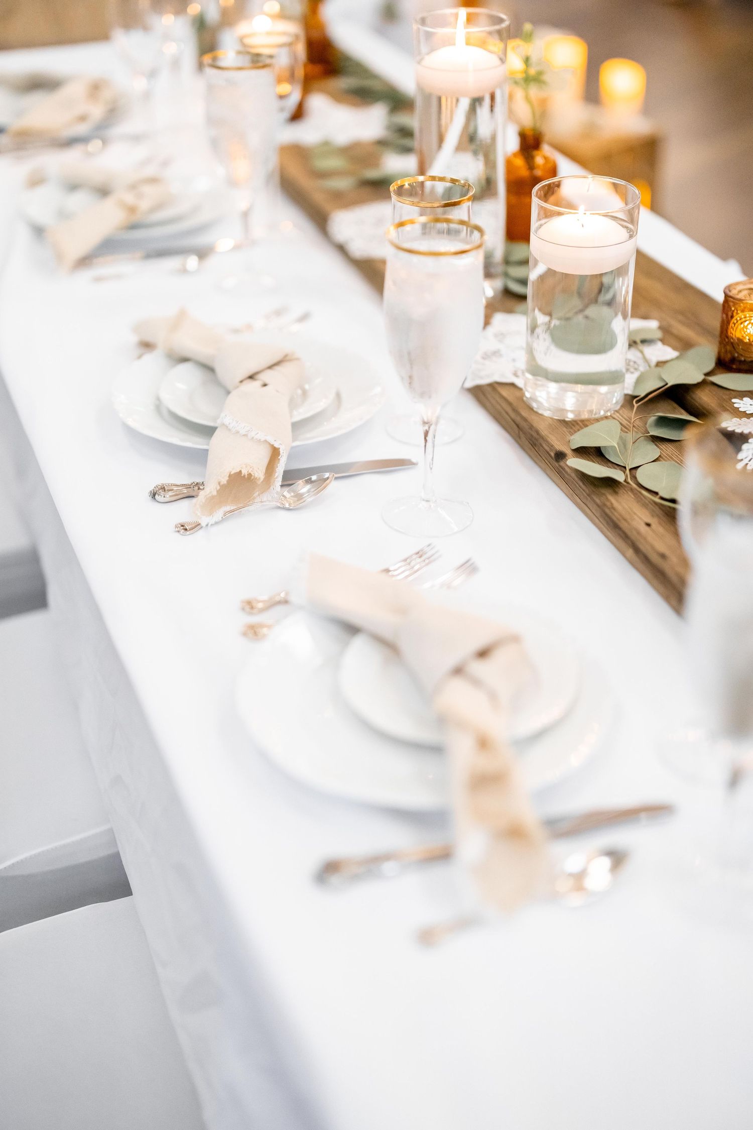 Wedding reception table featuring white plates, champagne napkins, gold-rimmed flutes, and glowing candle centerpieces.
