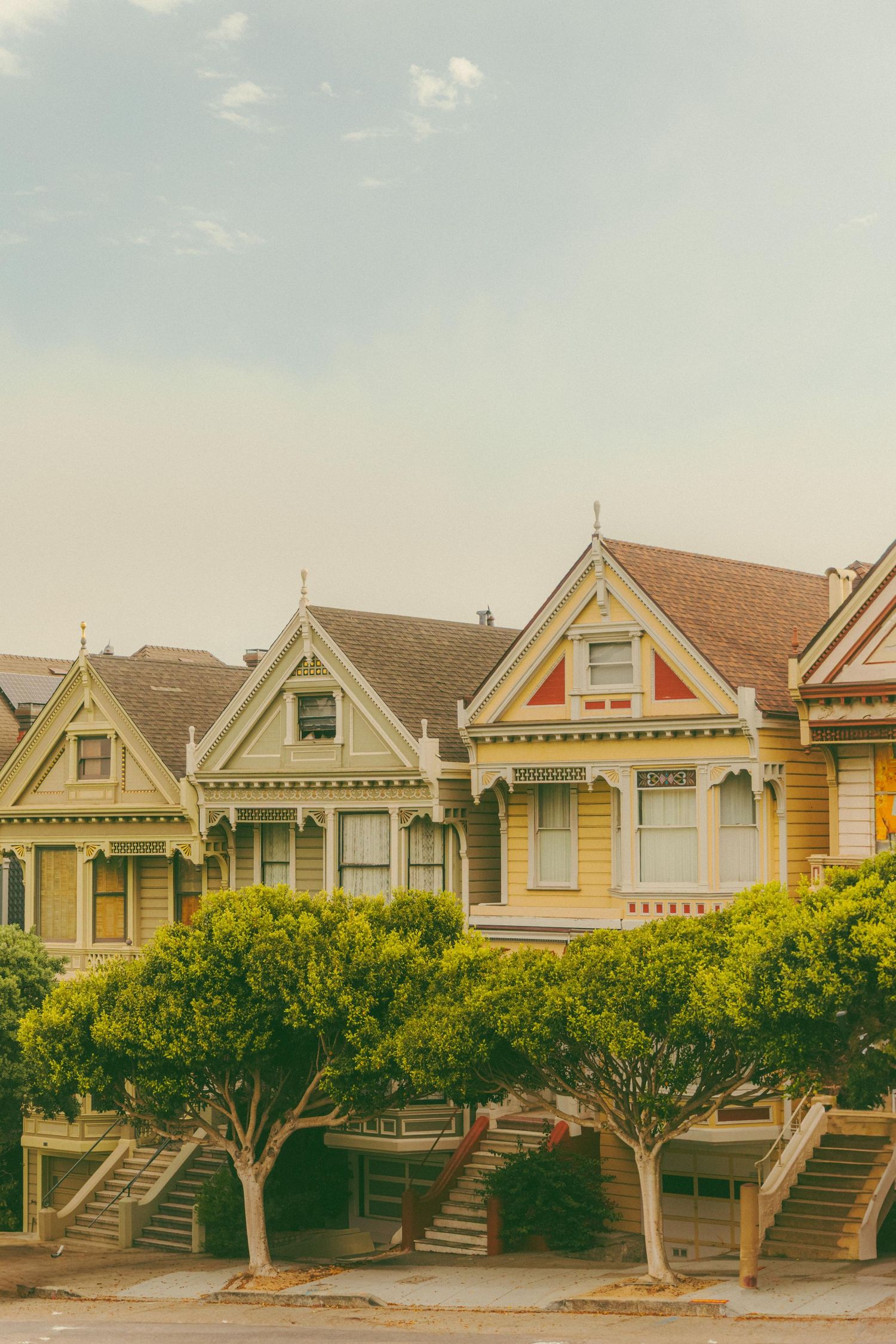Colorful Victorian houses known as the Painted Ladies line a San Francisco street, framed by green trees under a hazy sky.