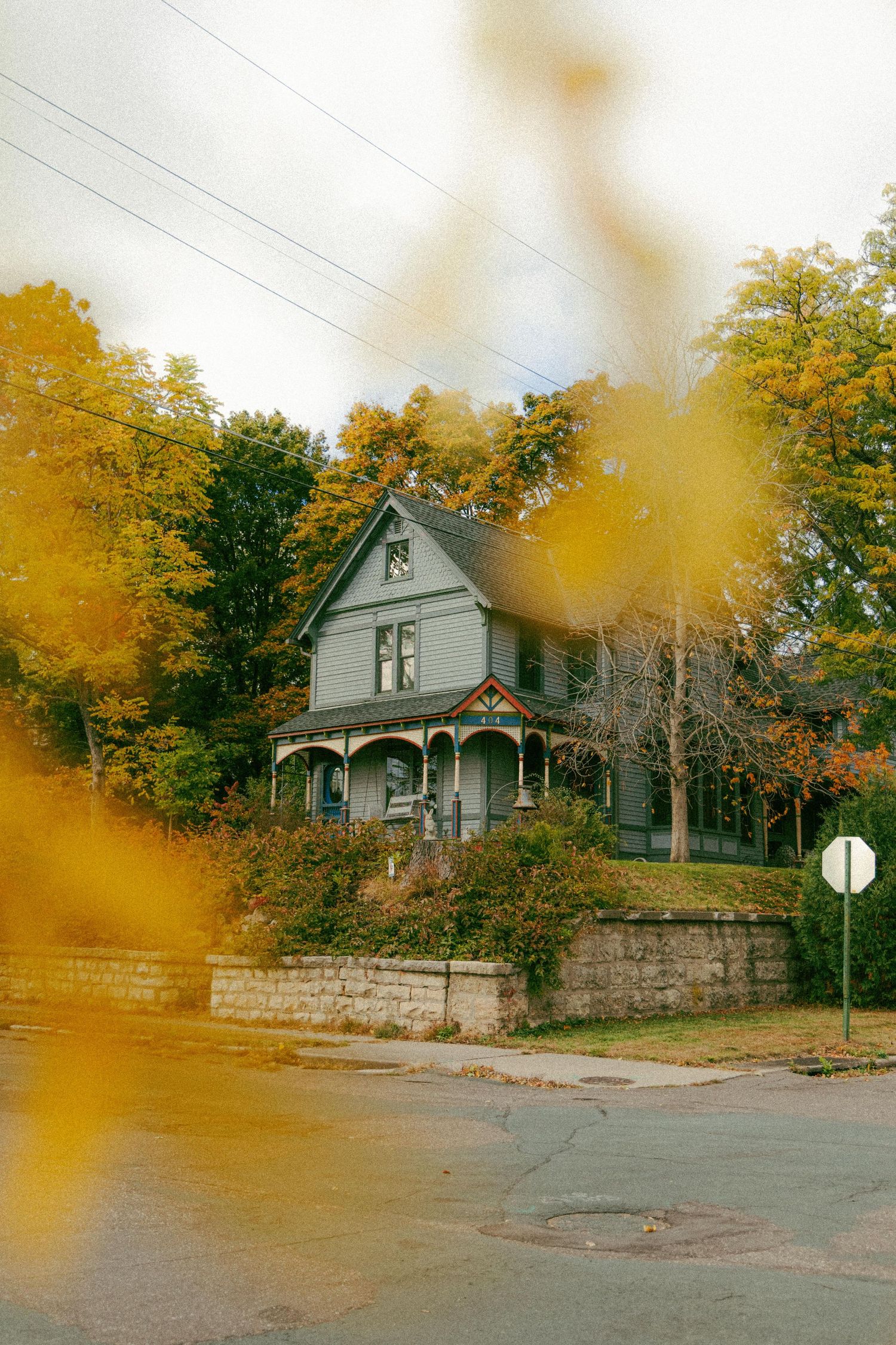 Historic gray Victorian house with wraparound porch surrounded by vibrant yellow autumn trees behind a stone retaining wall.