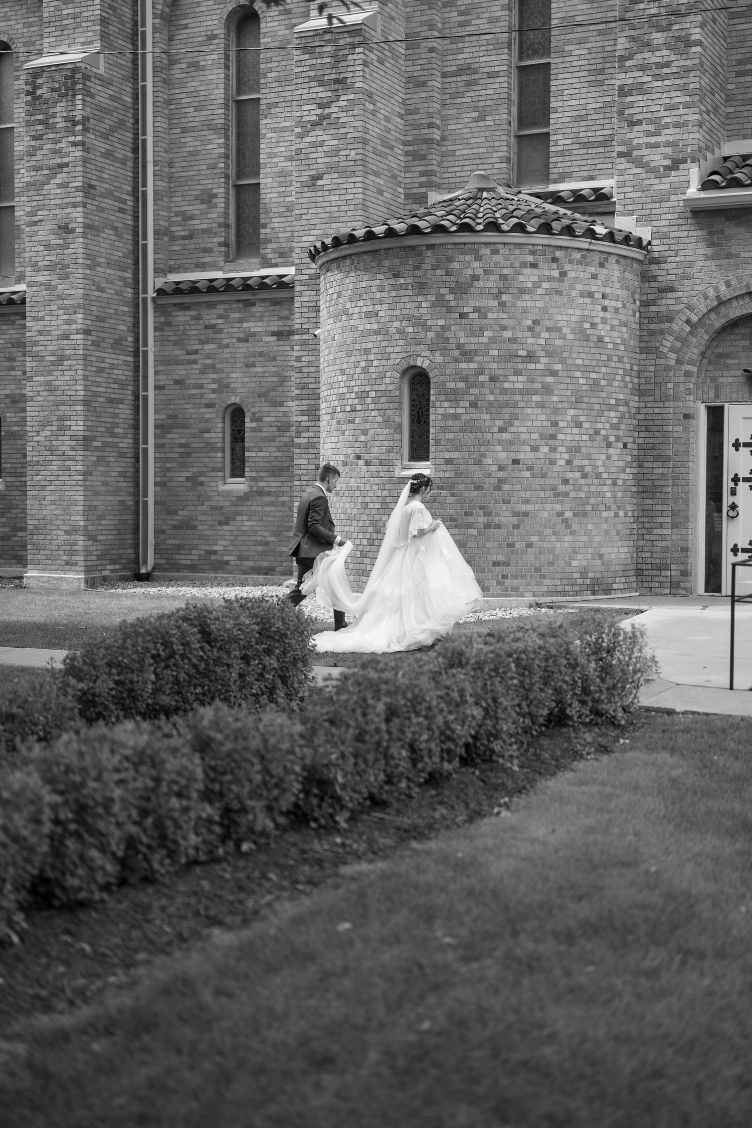 Black and white image of newlyweds strolling hand in hand outside a brick church.
