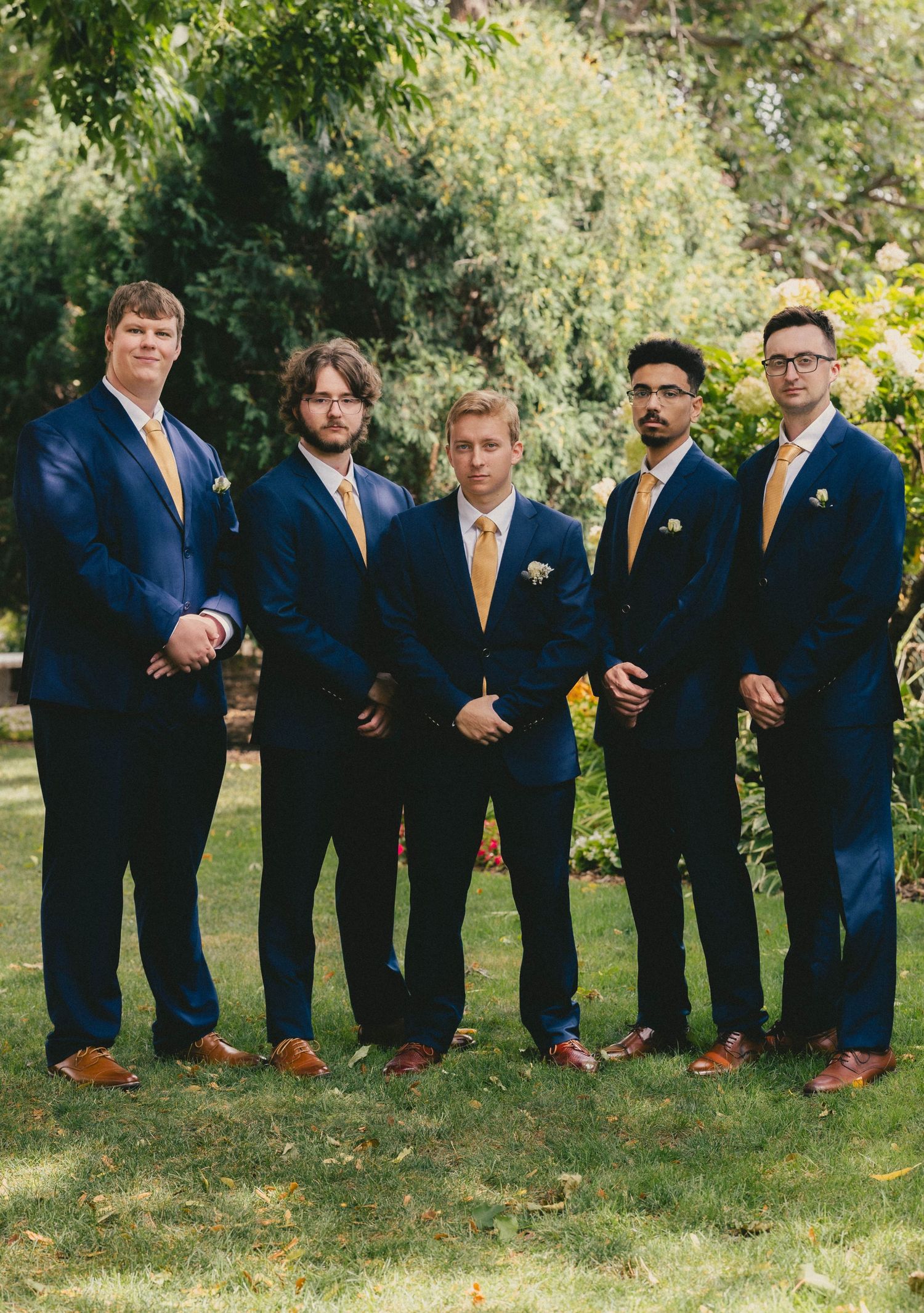 Five groomsmen in navy blue suits with gold ties pose outdoors on green grass for a wedding photo.