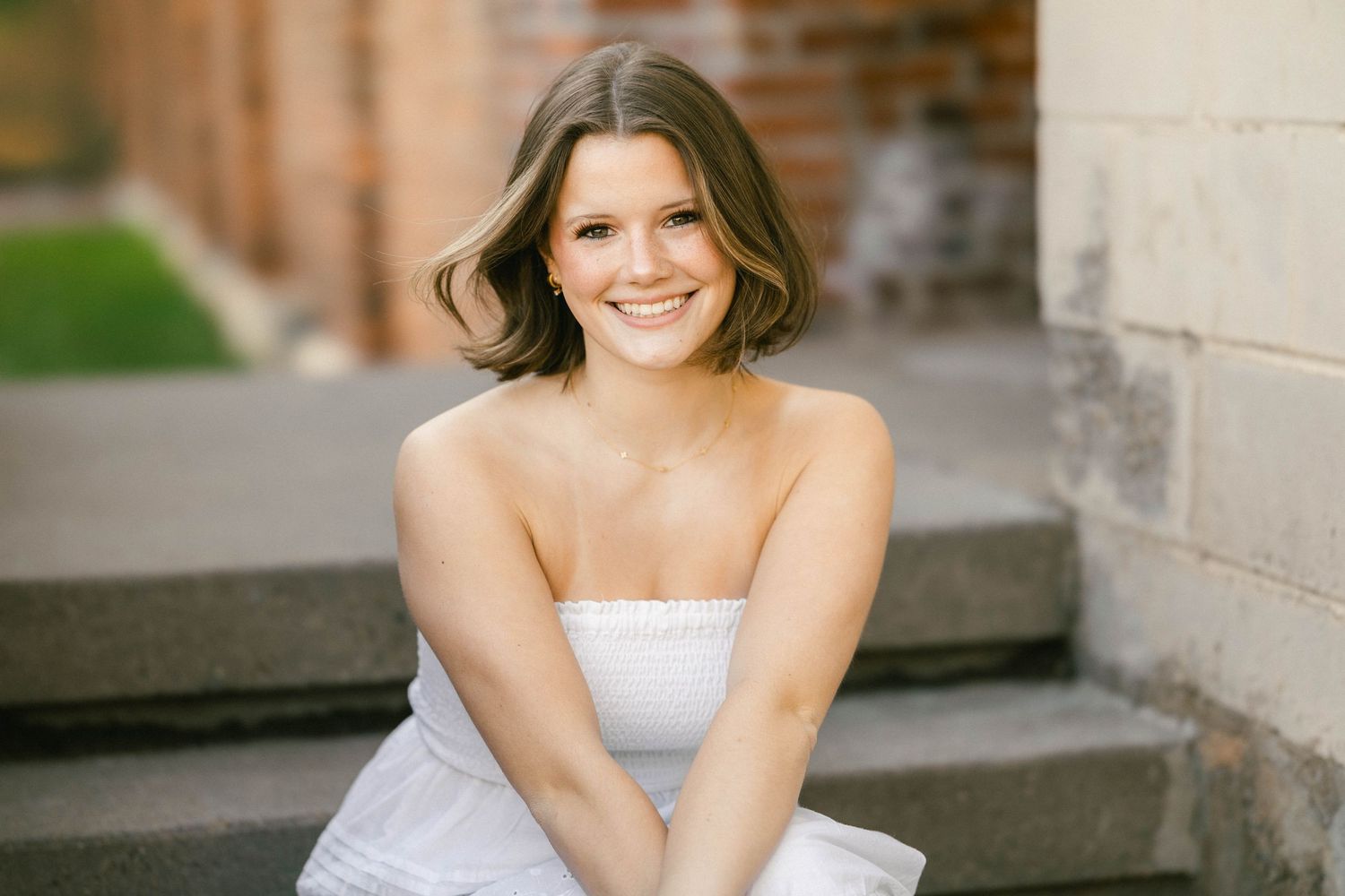 Smiling young woman in white strapless dress sitting on stone steps outdoors.