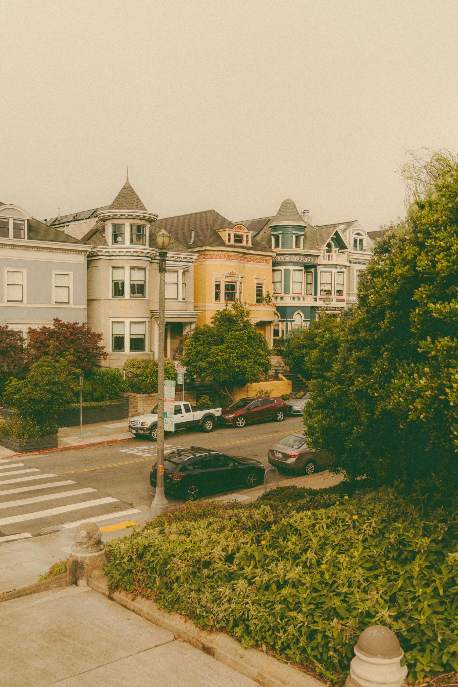 Row of colorful Victorian houses with turrets on a tree-lined San Francisco street with parked cars below.