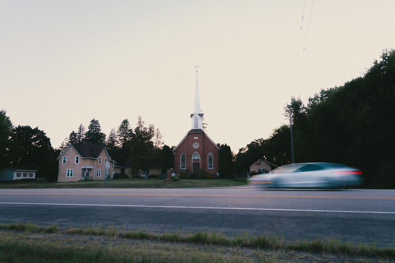 A car speeds past a red brick church with white steeple at a rural intersection surrounded by trees at dusk.